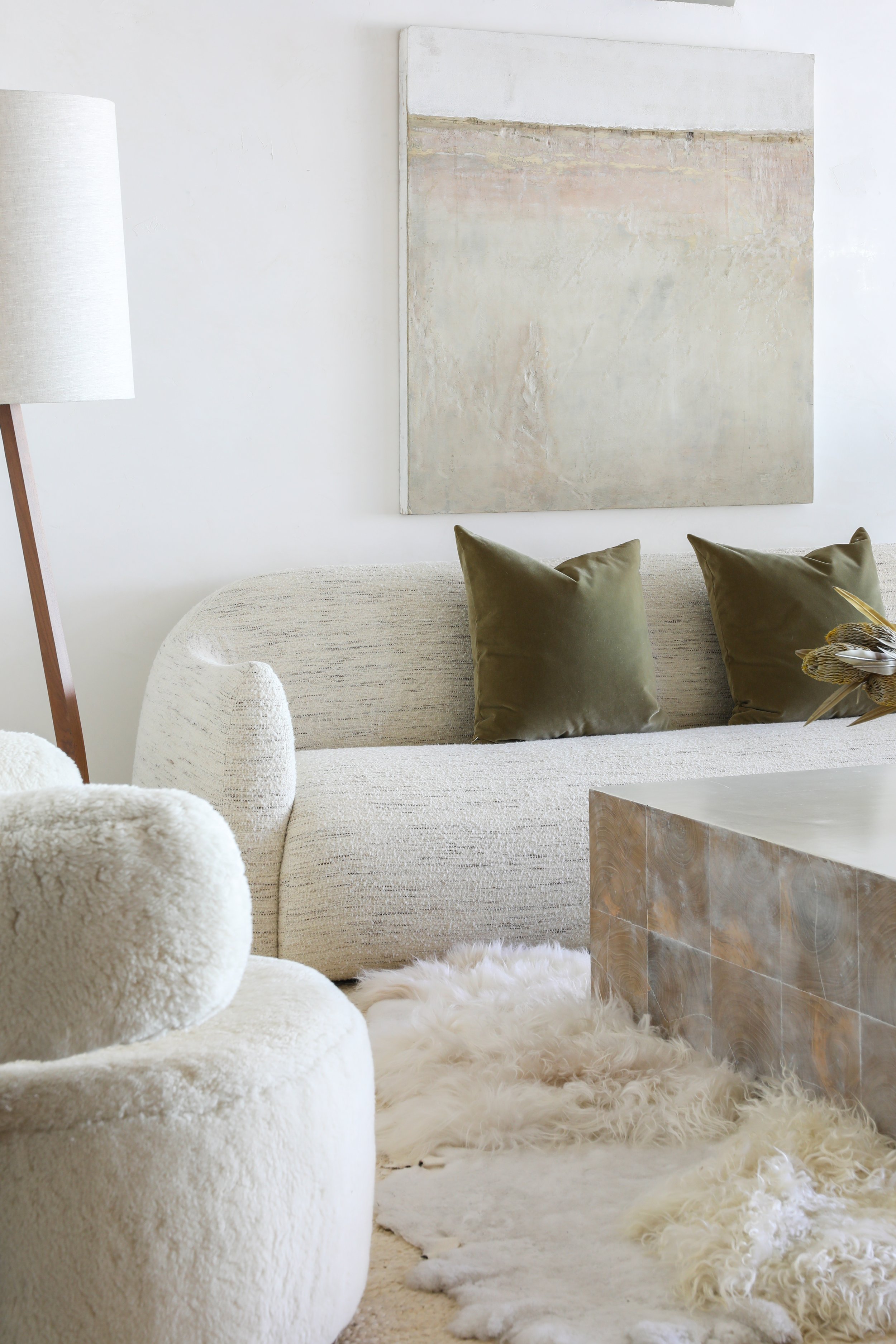 A modern, minimalistic living room with a beige sofa adorned with two green pillows, a fluffy white armchair, a textured wall art, a tall floor lamp, and a marble coffee table, with a white shaggy rug on the floor.