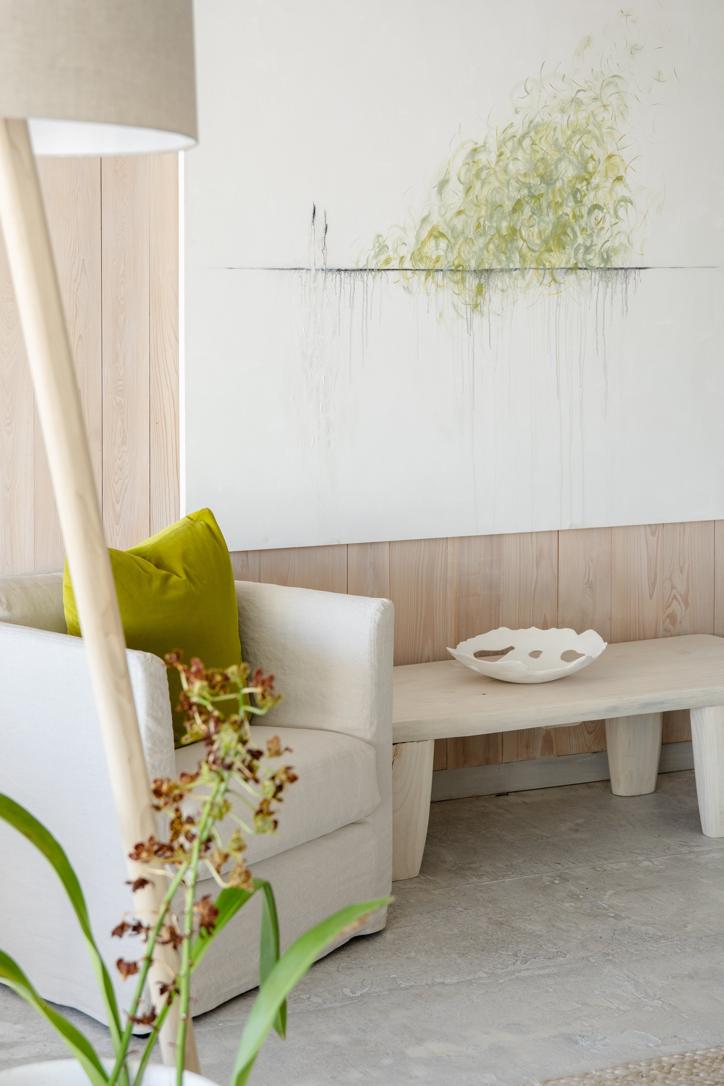 A living room corner with a white sofa, a green cushion, a light-colored wooden bench with a white ceramic bowl, a large abstract painting on the wall, a wooden panel wall, and a floor lamp.