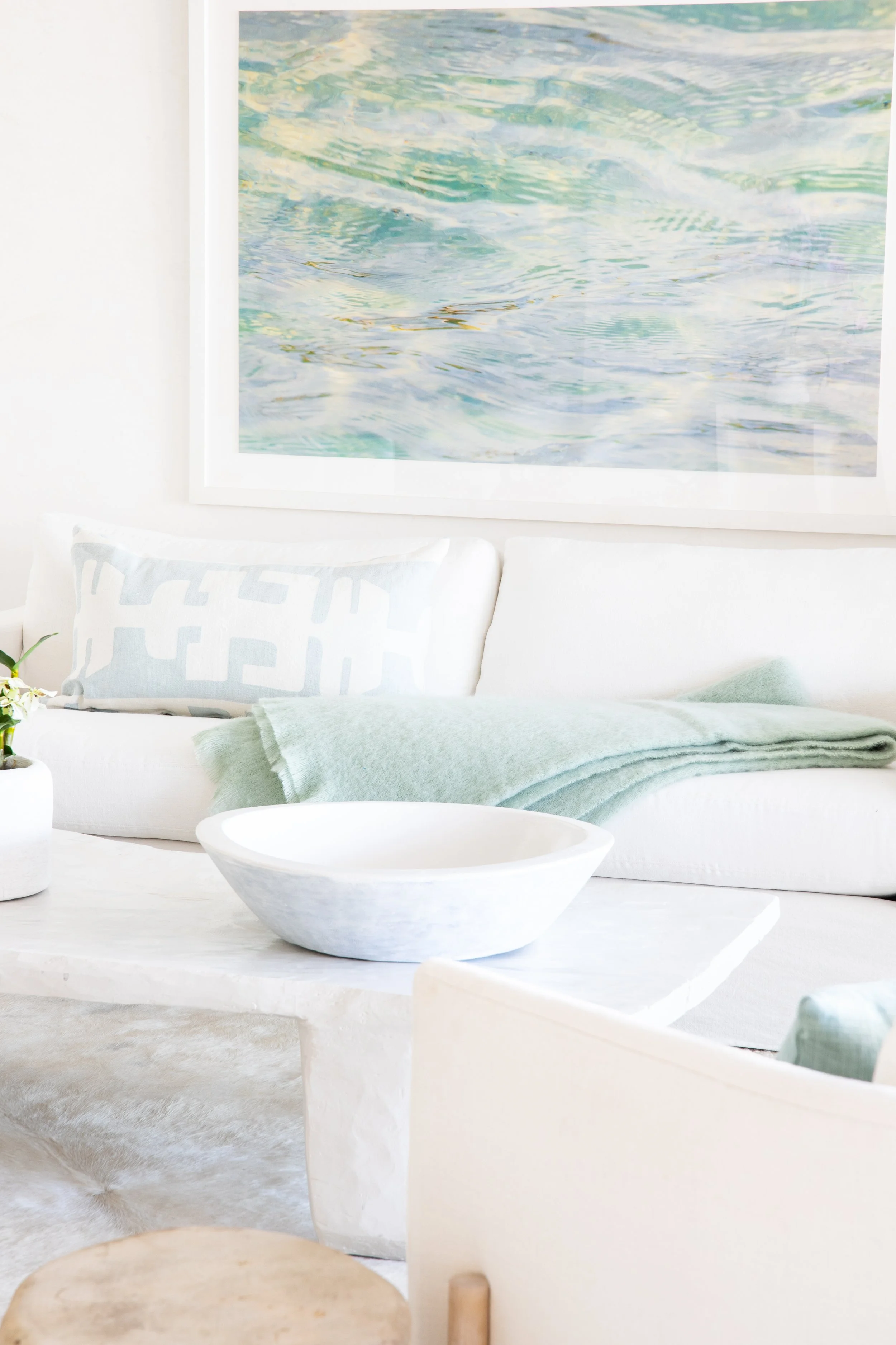 Living room with white sofa, light green blanket, patterned pillow, white coffee table with a shallow bowl, wall art of water, and a plant.