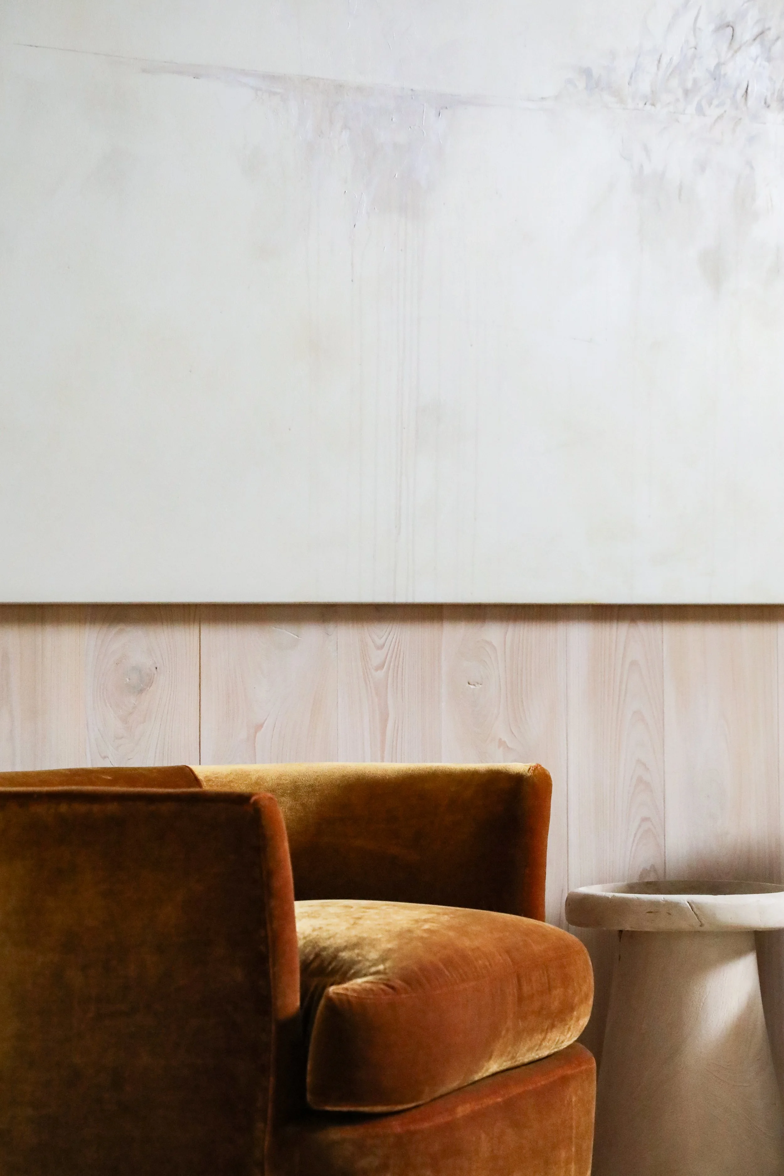 Close-up of a vintage brown velvet armchair and a small beige stool in front of a light wooden wall with an abstract painting above.