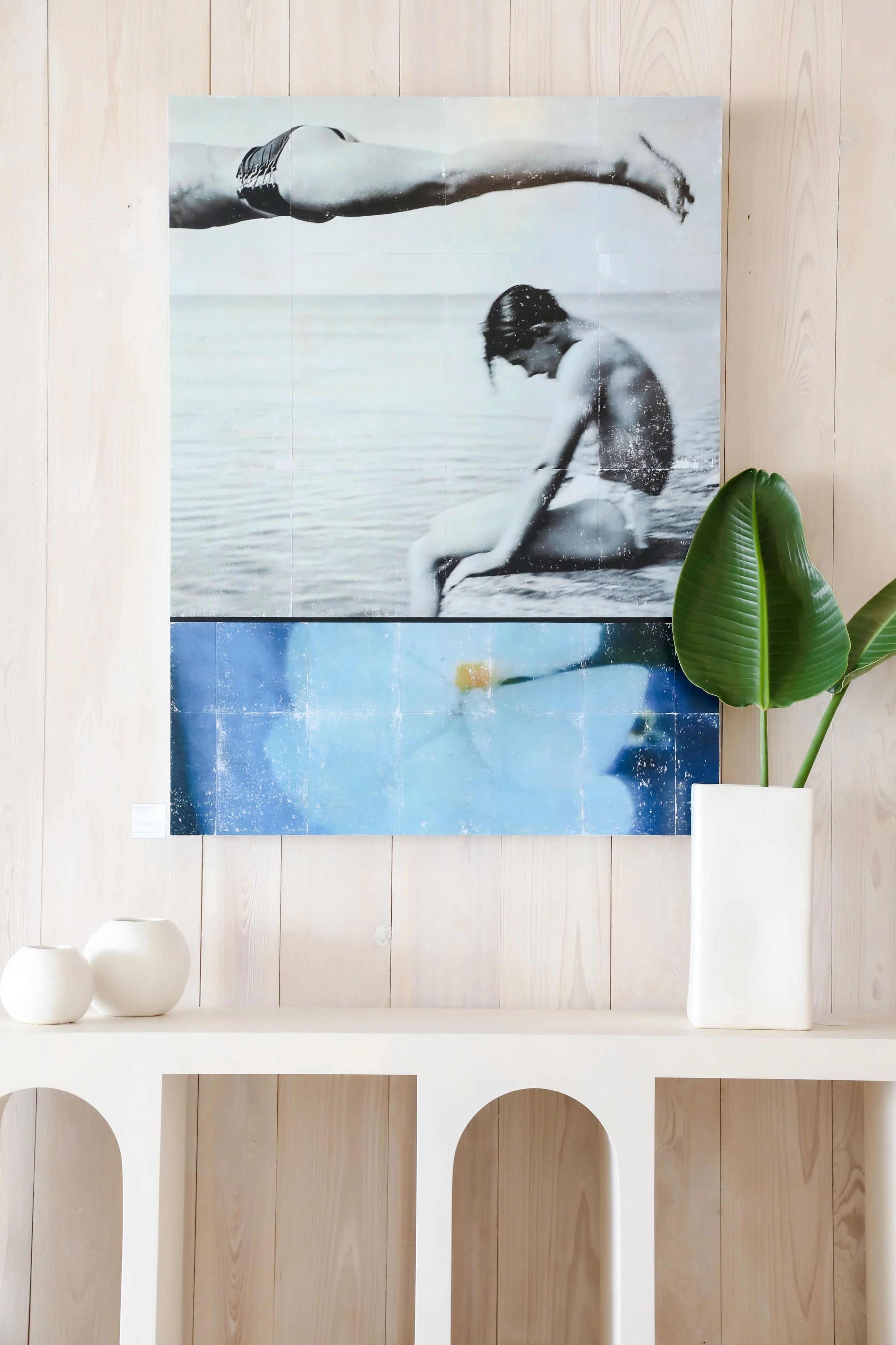 A black and white photograph of a person sitting at the edge of a body of water, with another person jumping into the water, mounted on a wooden wall with a plant and white decorative objects below.