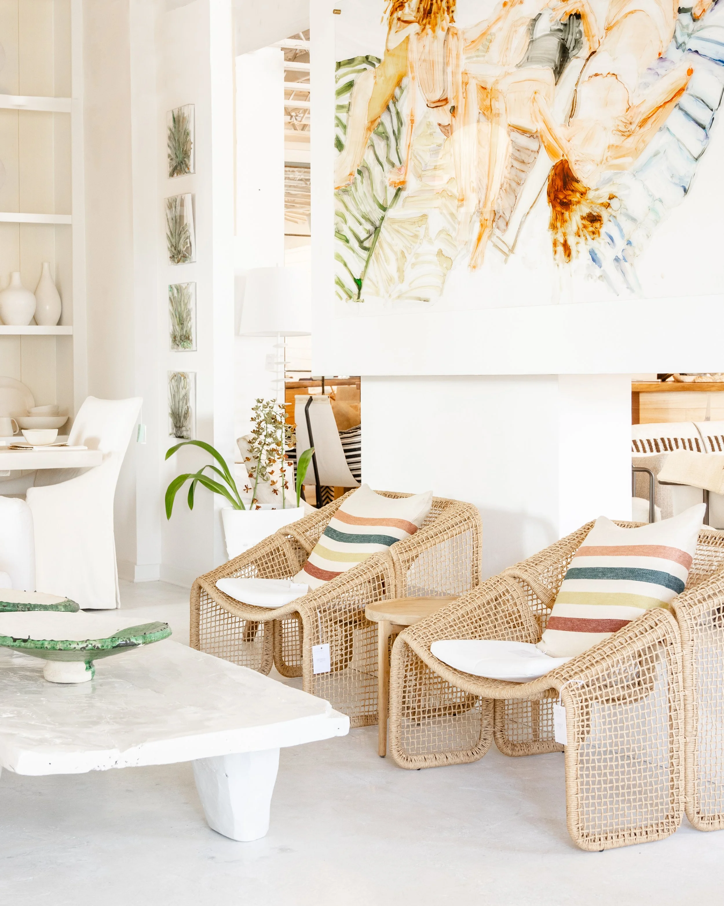 Bright, airy interior with woven wicker chairs, striped pillows, abstract wall art, potted plants, and a rustic white coffee table.