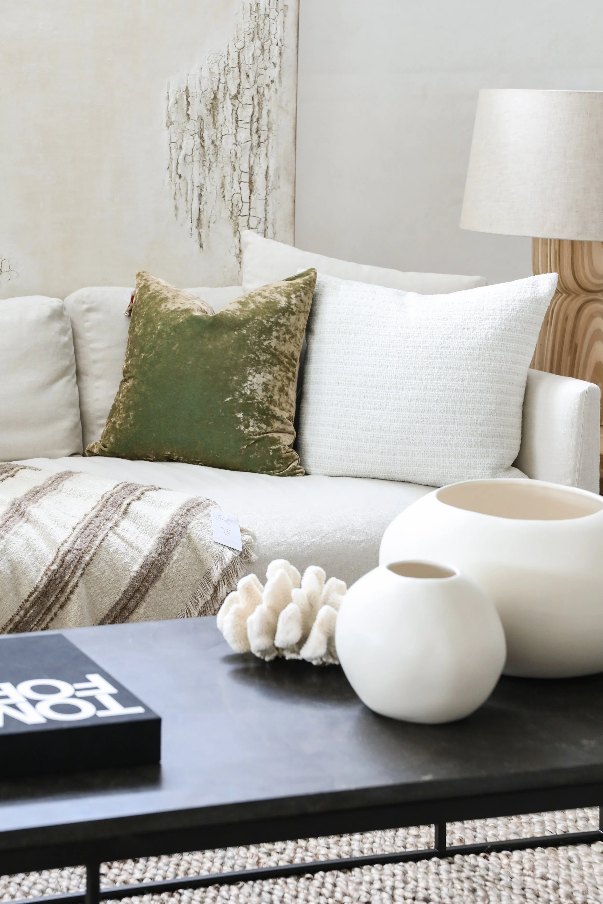 Living room with white sofa, green and white pillows, a beige blanket, a white textured lamp, and white decorative vases on a black table.