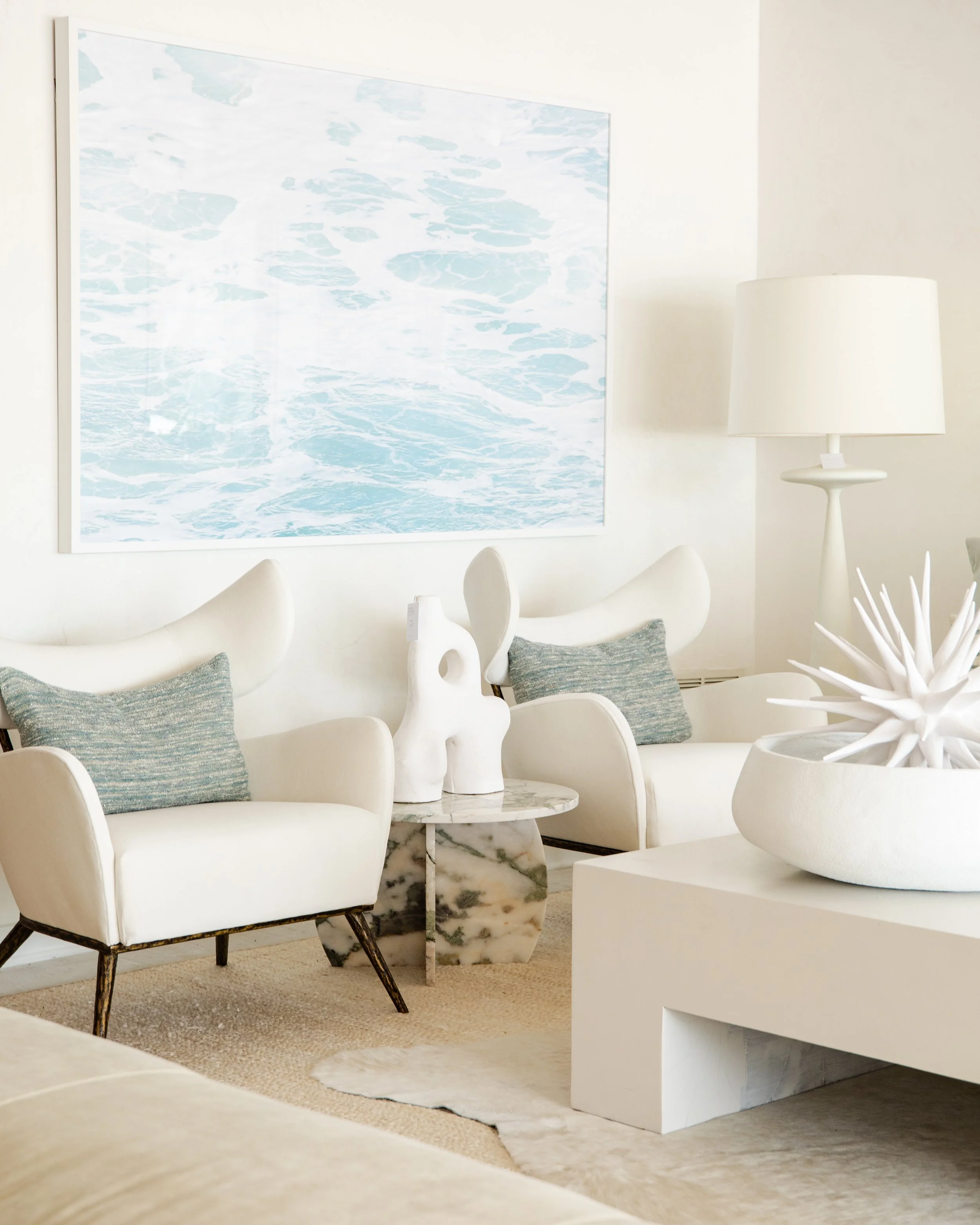 Modern living room with white armchairs, blue pillows, marble side table, abstract ocean-themed artwork, white lamp, and decorative sculpture.