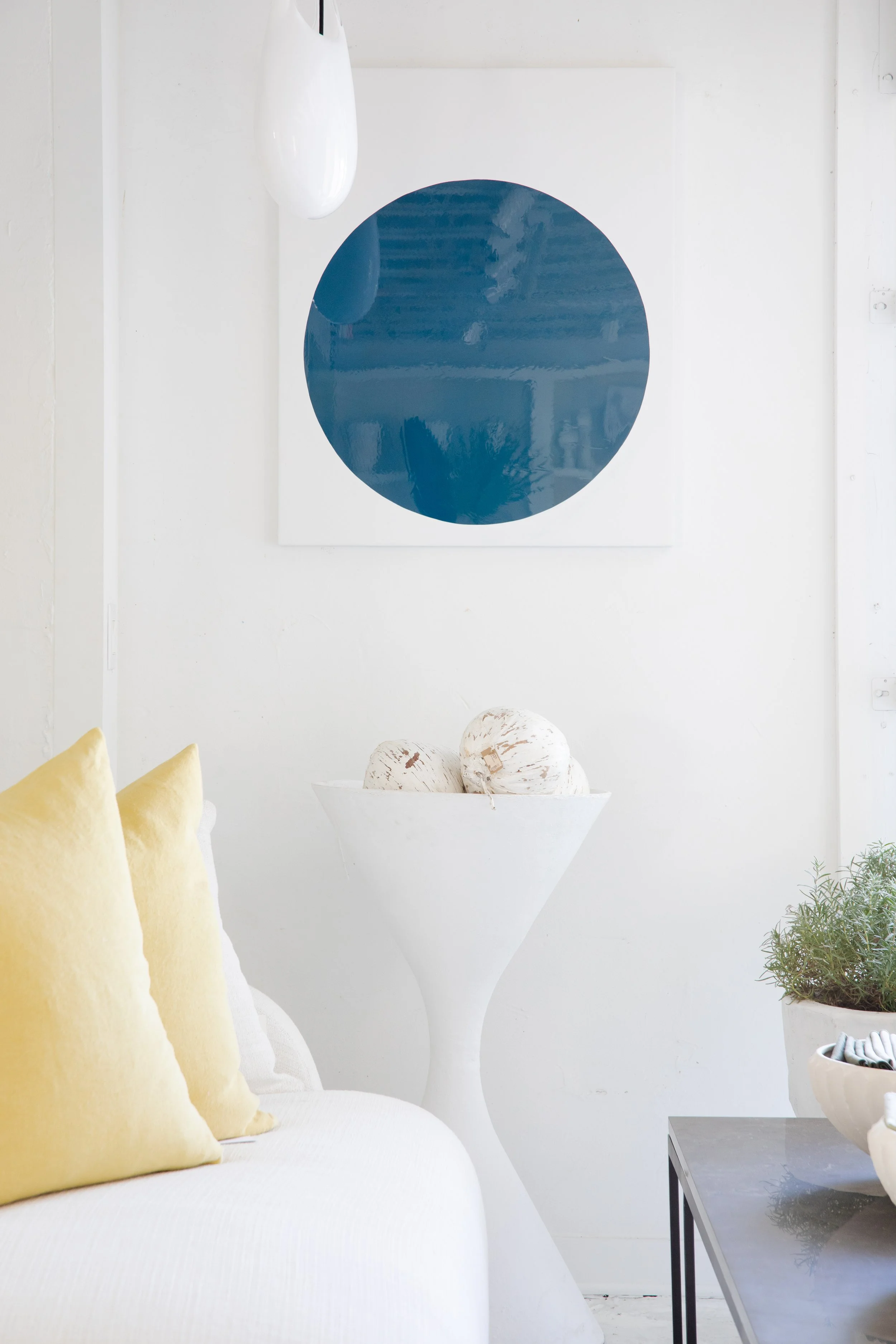 A white living room corner with yellow pillows on a white sofa, a tall white vase with three spherical decorative objects, a modern black table, a green plant in a white pot, and a minimalist abstract artwork with a blue circle on a white background.