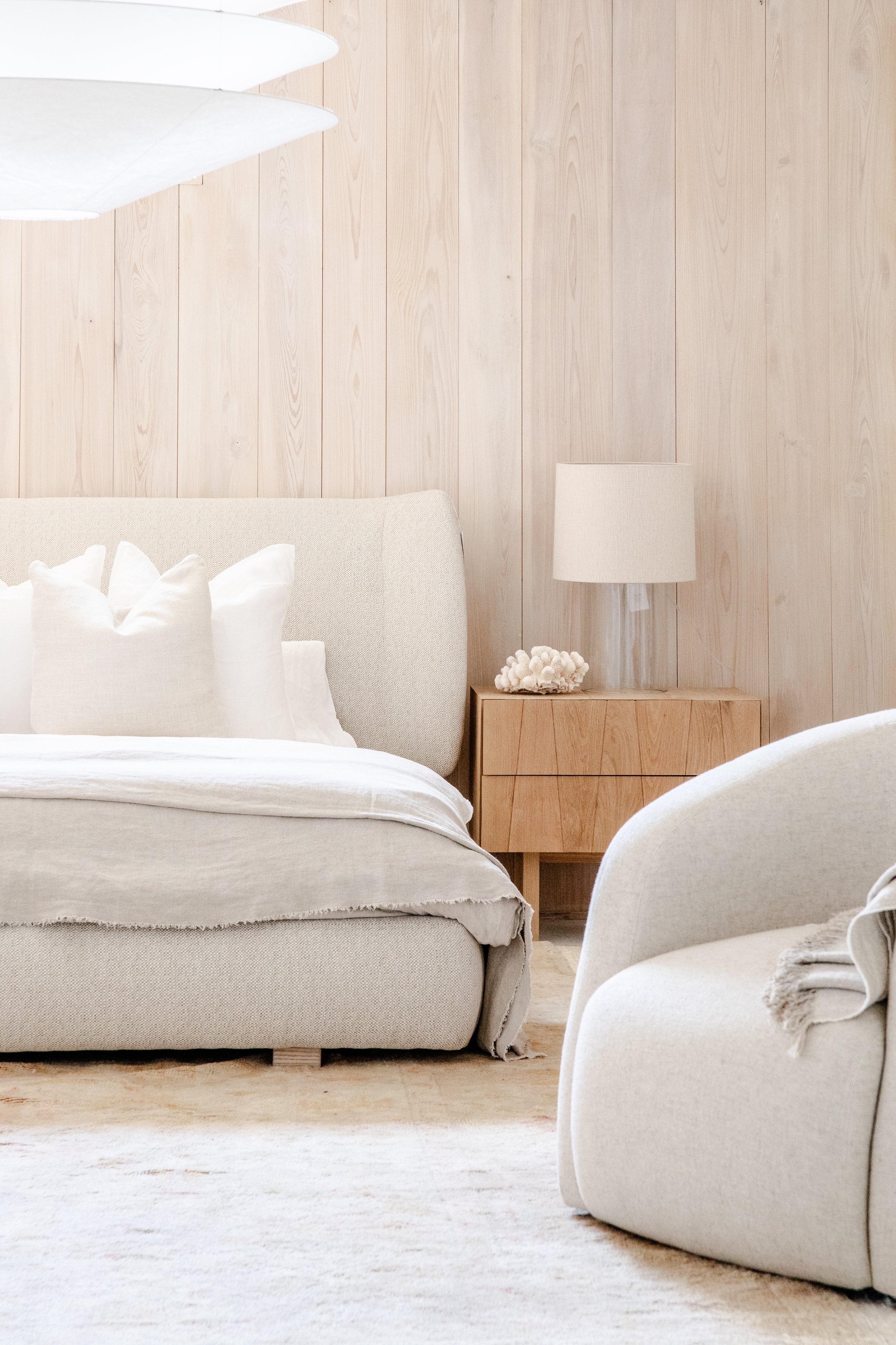 A cozy bedroom with a cream upholstered bed, white pillows, a light wood nightstand with a white lamp, and beige wooden paneling on the wall.
