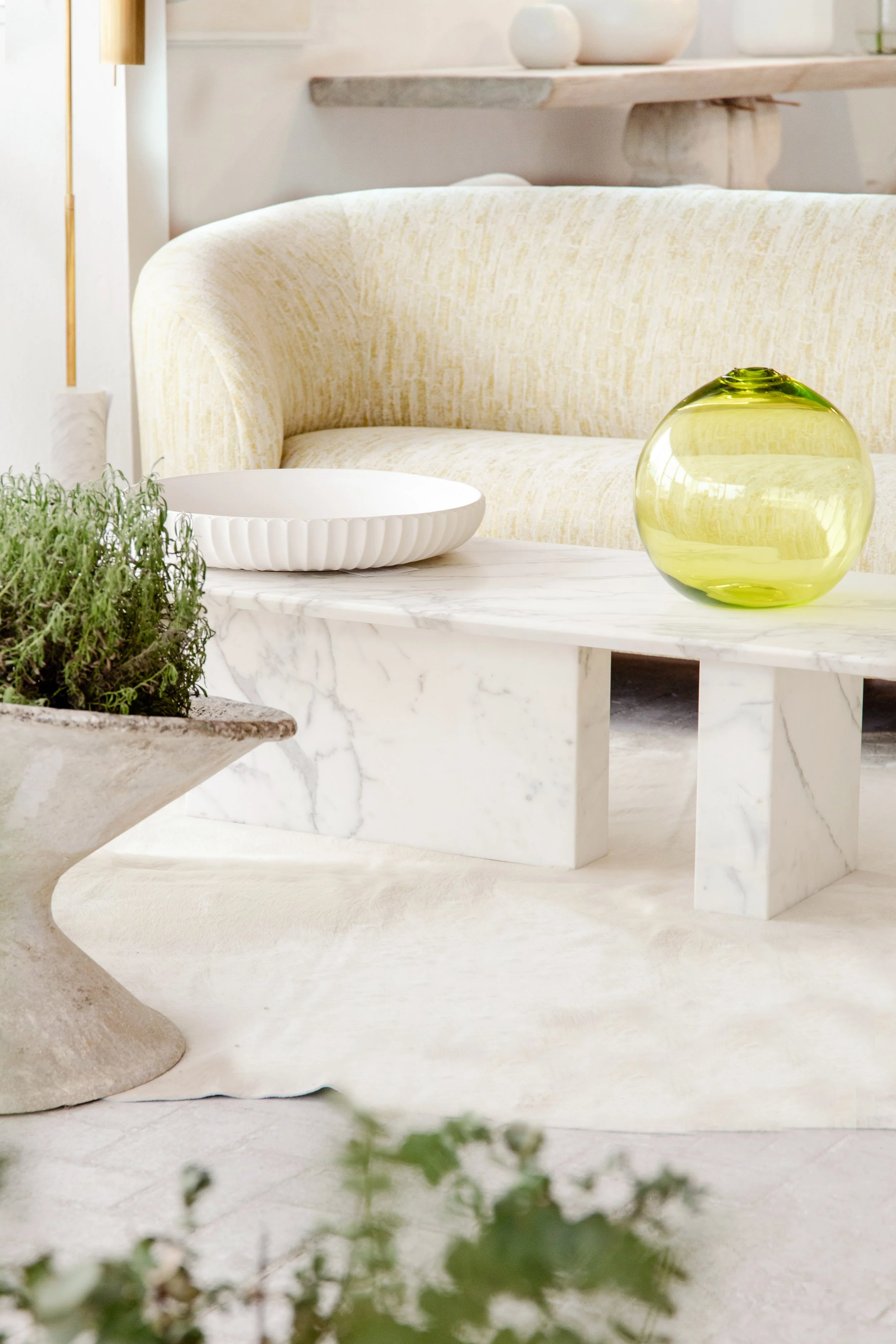 A living room with a white marble coffee table, a beige sofa with a yellowish pattern, a white dish, and a green glass vase on the table. A potted plant is on a side table, and a shelf with white decorative objects is in the background.