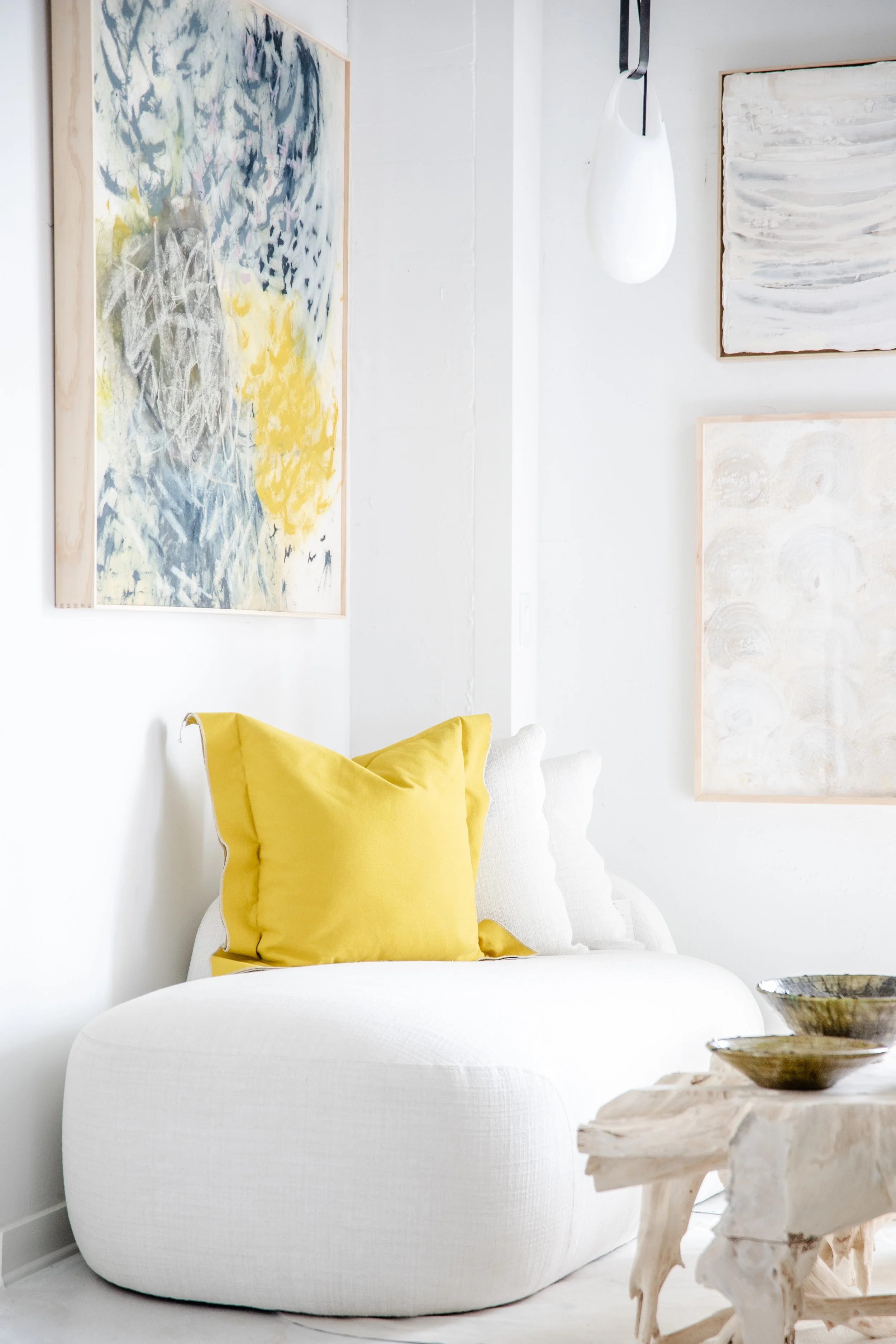 A bright room with white walls and contemporary decor, featuring a white rounded sofa adorned with a yellow pillow, next to a small, natural wooden side table with decorative bowls. Abstract paintings in soft colors hang on the walls.