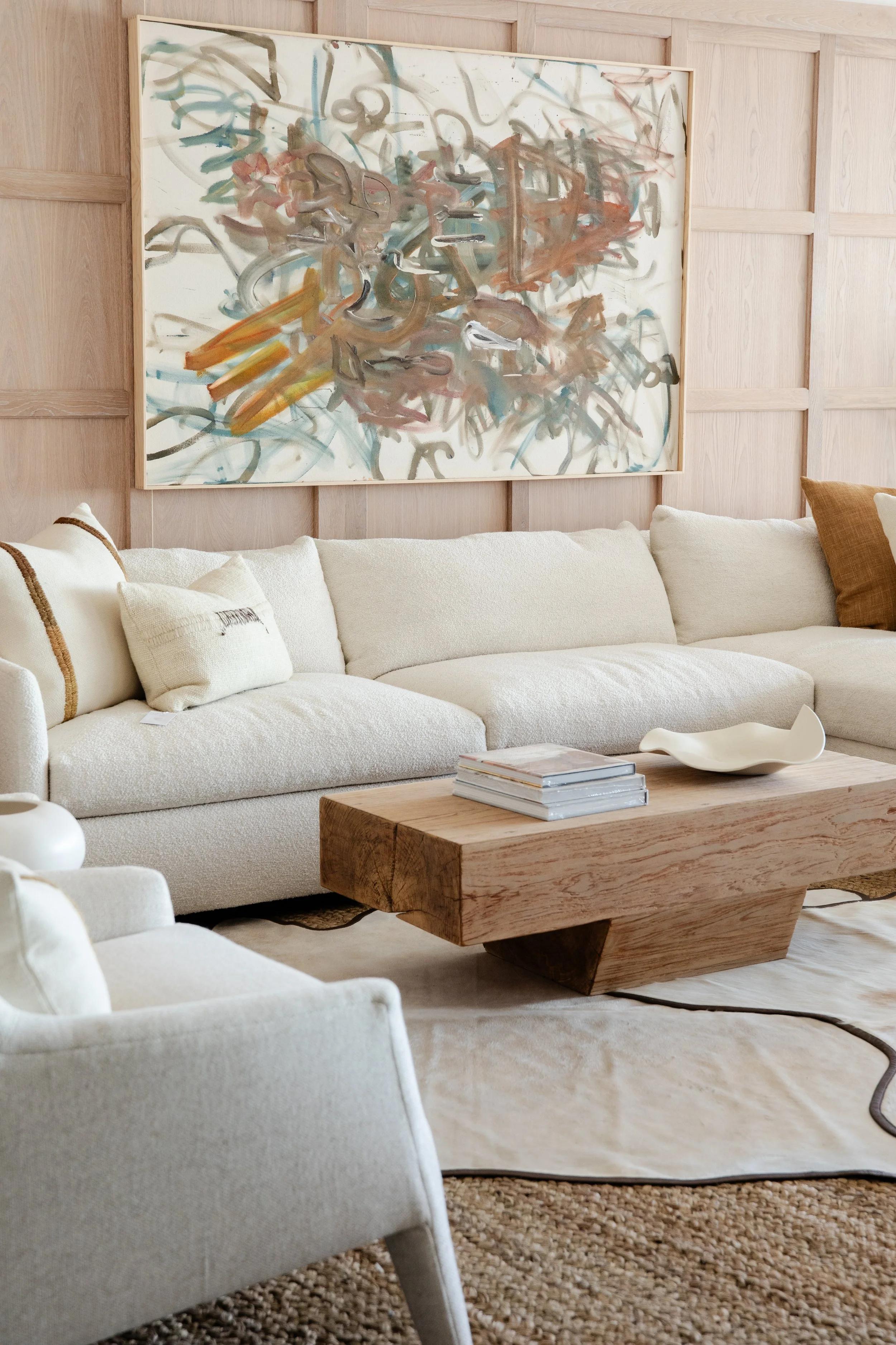 Living room with cream-colored sofa, abstract painting on wooden-paneled wall, wooden coffee table with books and decorative bowl, neutral-toned pillows and rug.