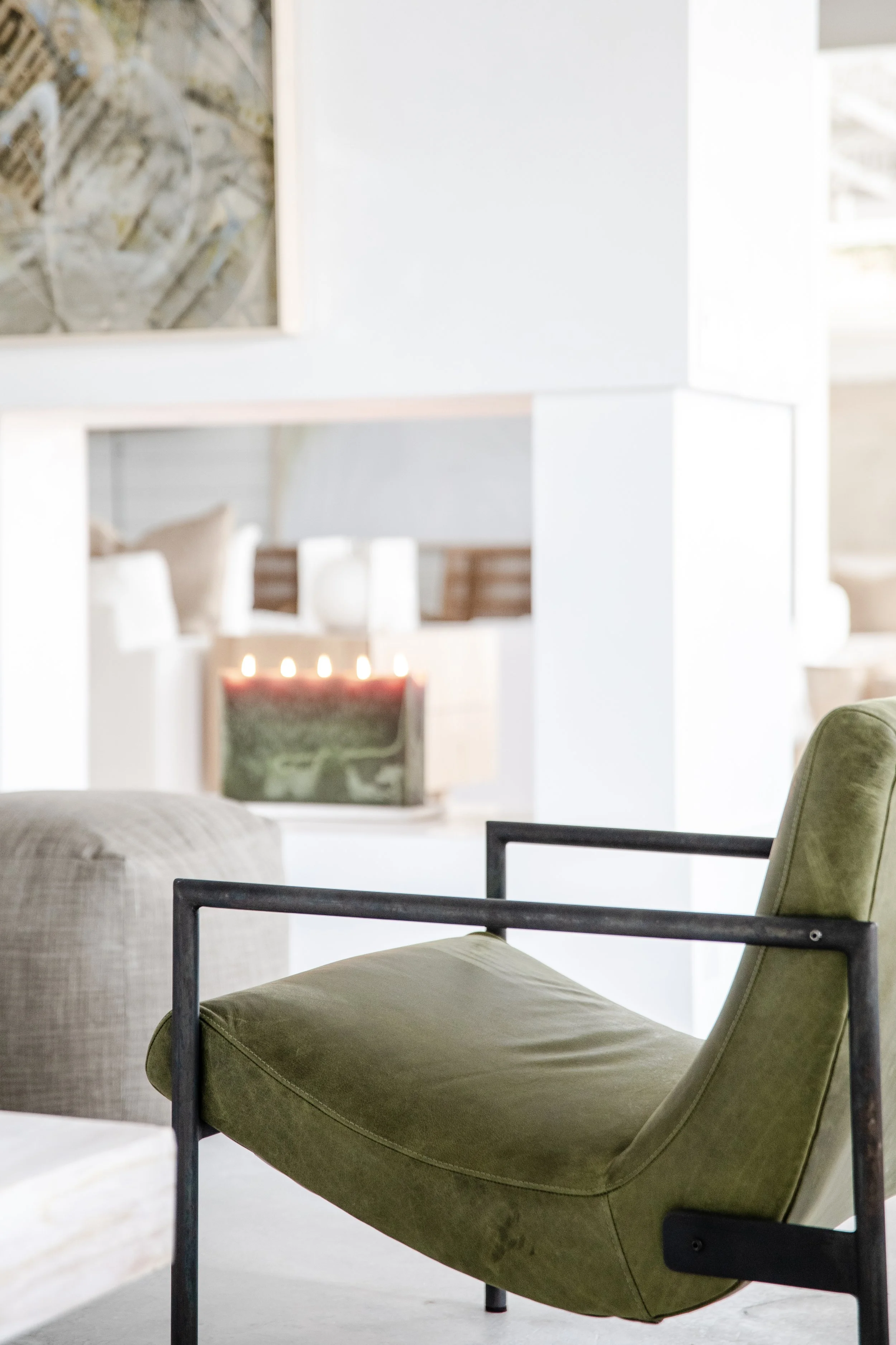 A modern living room with a green upholstered lounge chair with black metal frame, white walls, a beige sofa, and a fireplace with lit candles in the background.