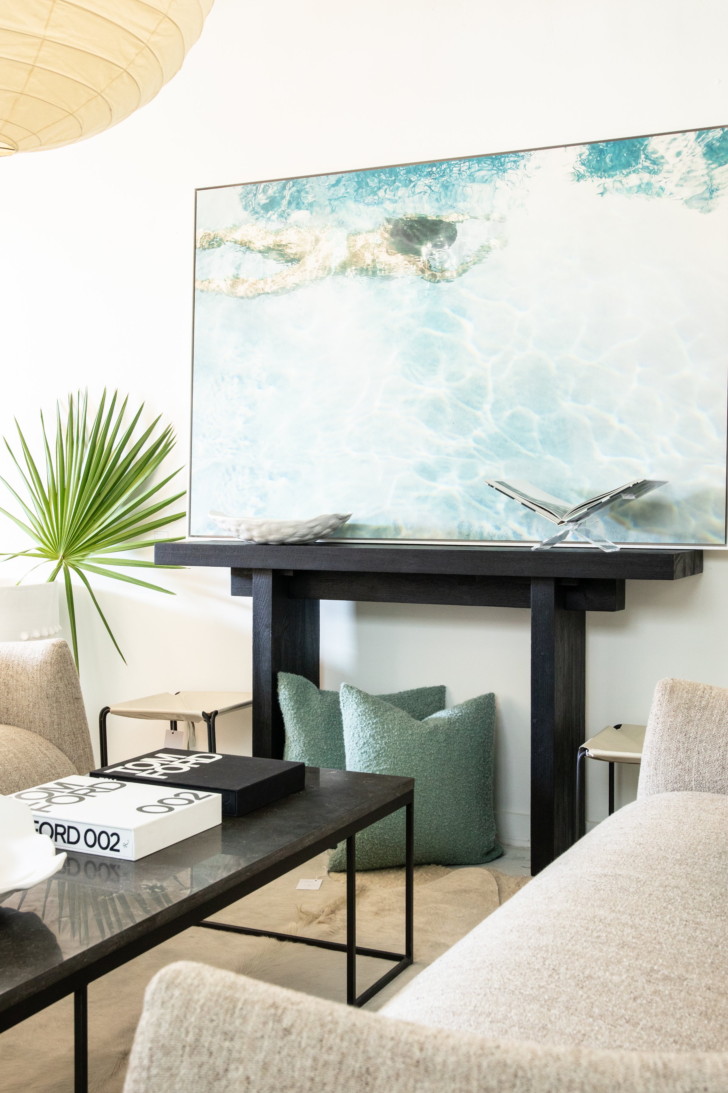 Living room with a large artwork of a swimming pool on the wall, a black console table underneath, a green textured pillow on the floor, books and a book stand on the table, and a beige sofa with a black side table in the foreground.