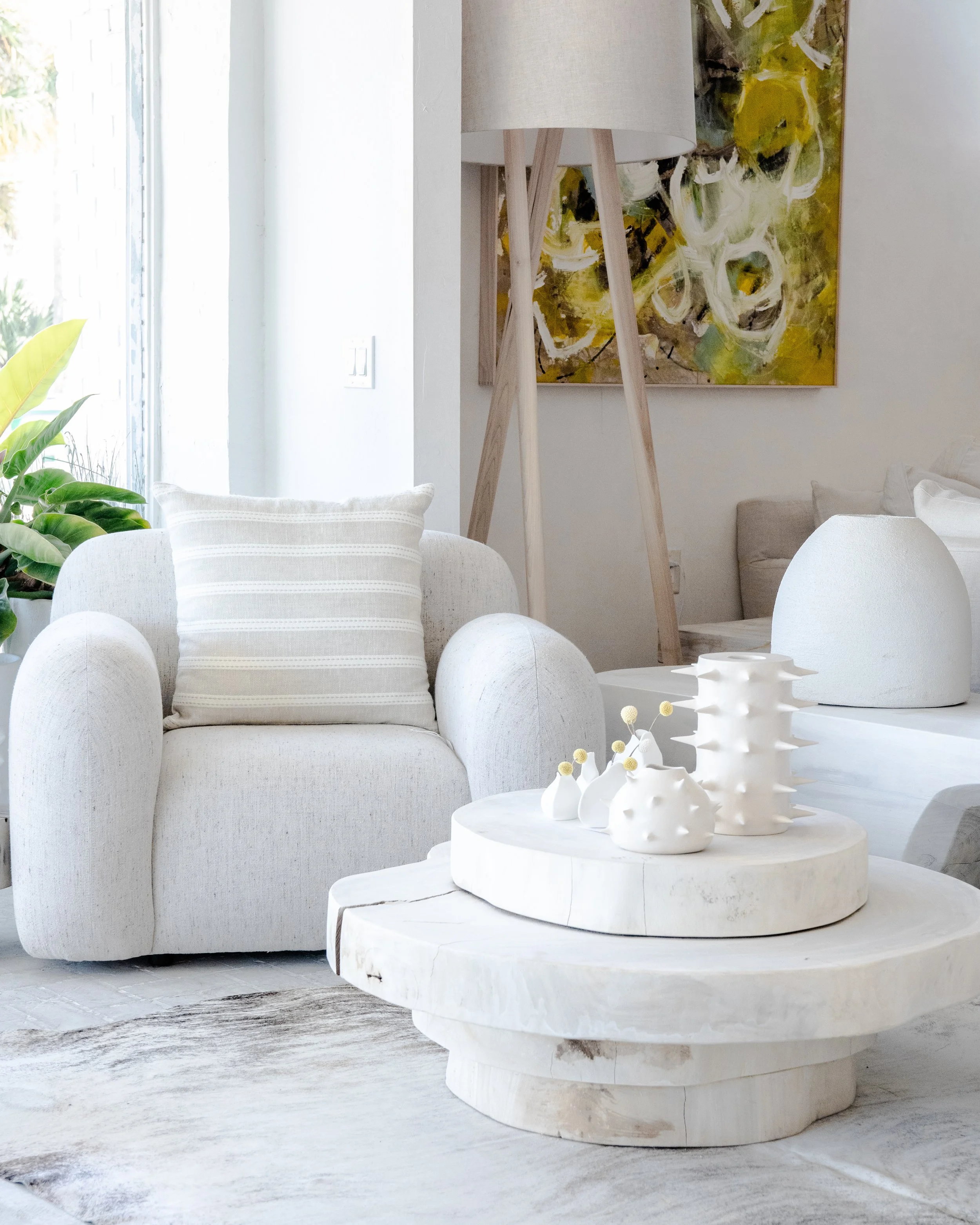 A modern living room with white and beige furniture. There is a beige armchair with a striped pillow, a white marble coffee table with white ceramic vases, a beige floor lamp, a large abstract yellow and black painting, and a potted plant near a larg