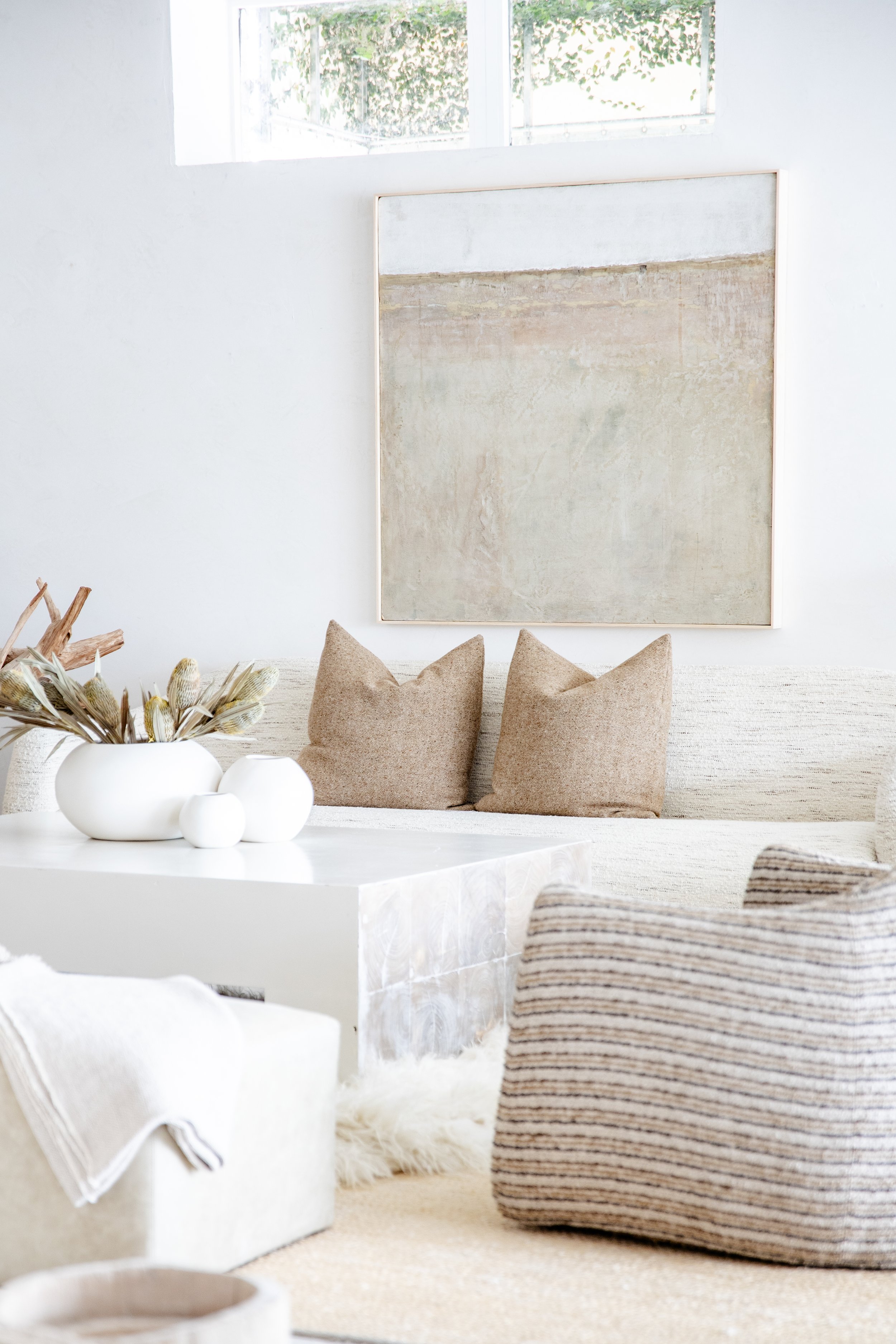 Minimalist living room with a neutral color palette, featuring a beige sofa with matching pillows, a white marble coffee table with white decorative vases, a textured striped armchair, a cream rug, and an abstract artwork hanging on the wall beneath 
