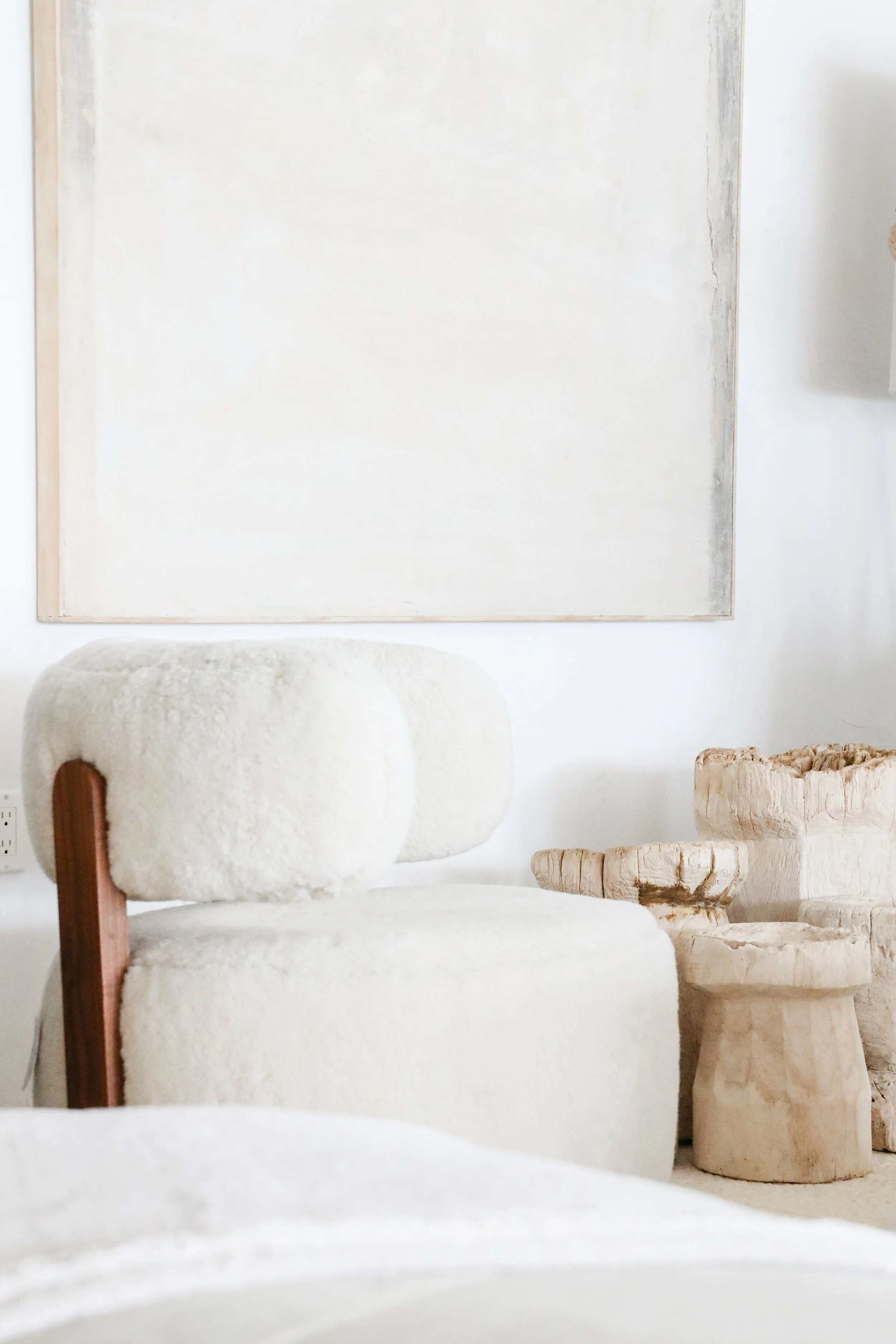 Minimalist interior with a white textured armchair, two wooden carved stools, a large empty canvas on the wall, and soft natural light.