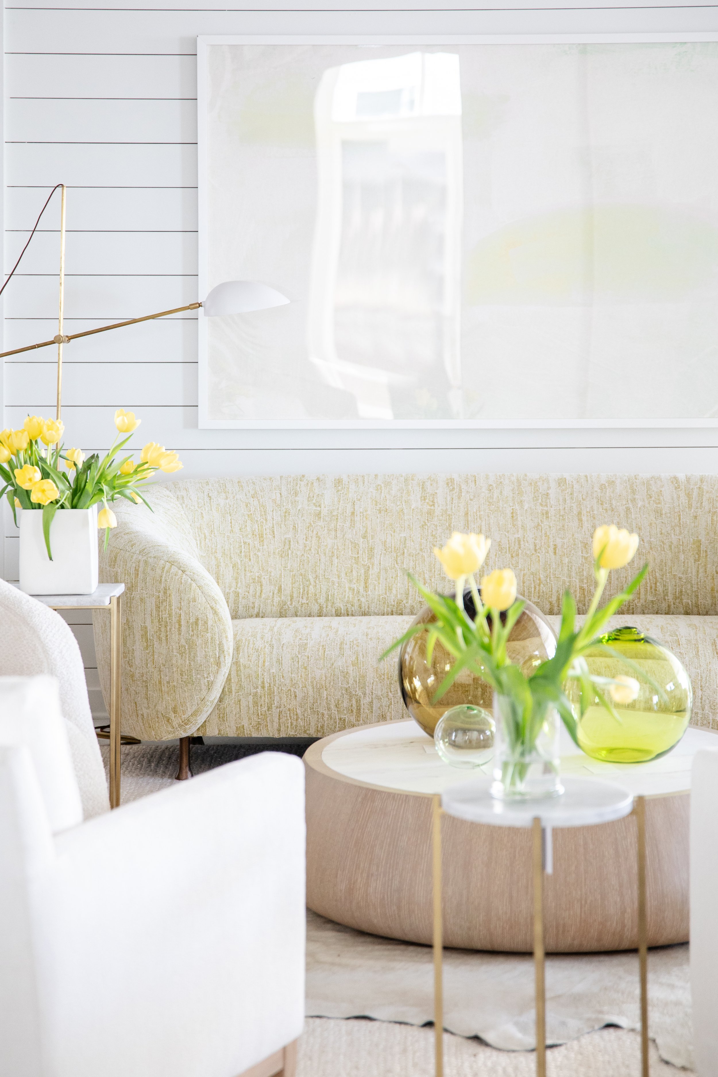 Bright living room with yellow tulips in vases on a white and gold coffee table, beige sofa, and white walls with artwork.