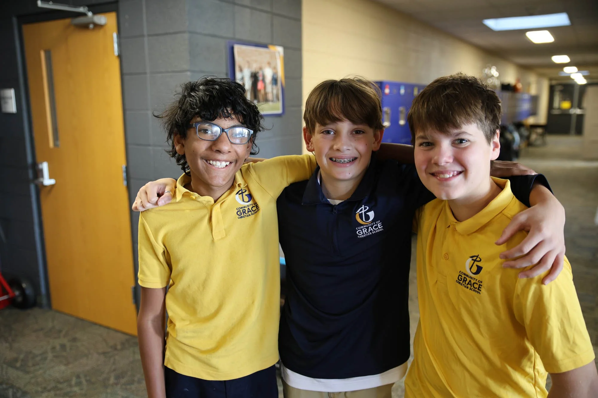 a group of three middle school friends with arms around each others shoulders and wearing Community of Grace Christian School branded polo shirts in a school hallway