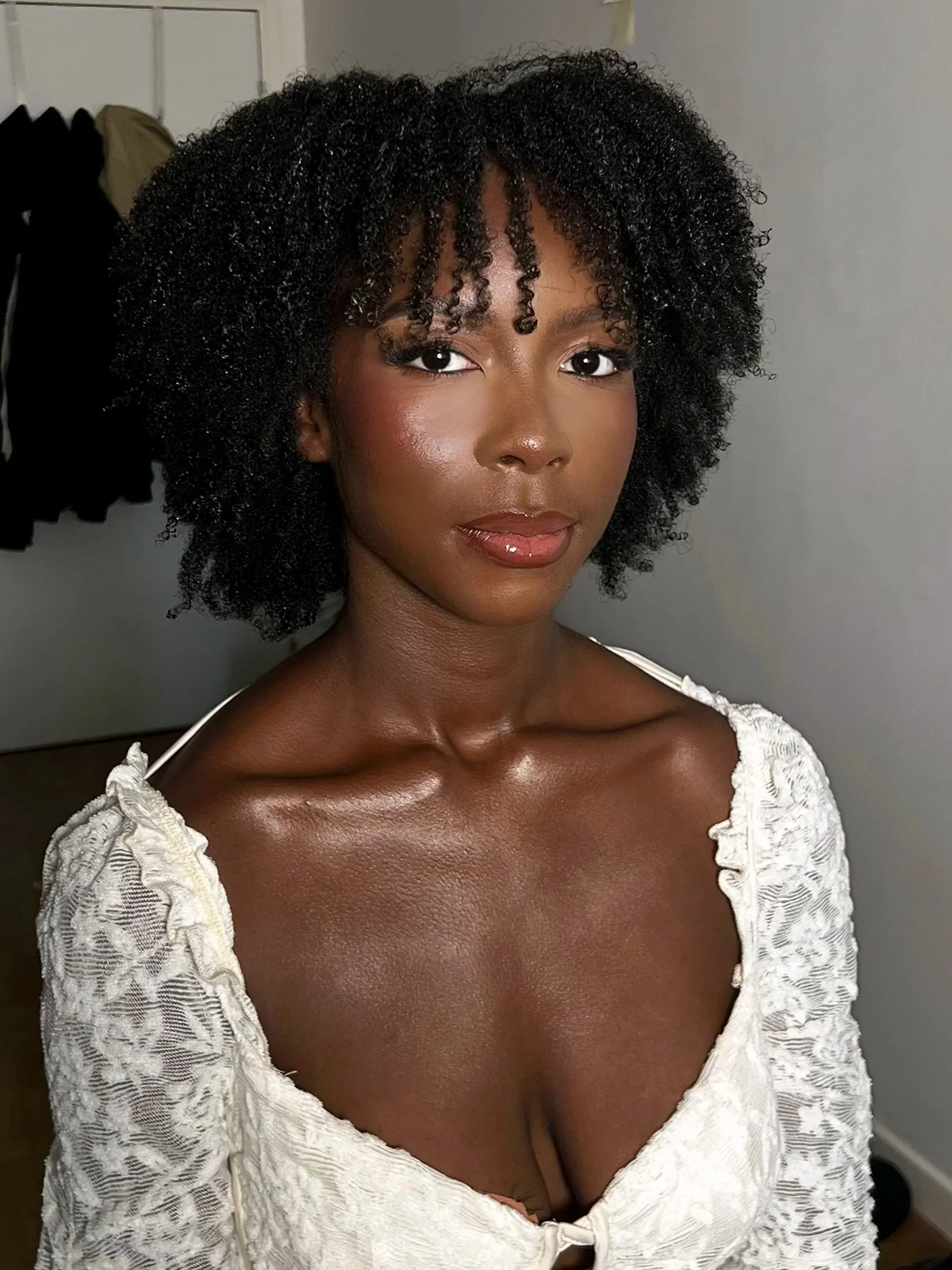 Une femme noire avec des cheveux bouclés courts, portant une robe blanche en dentelle, se tenant dans une pièce intérieure.