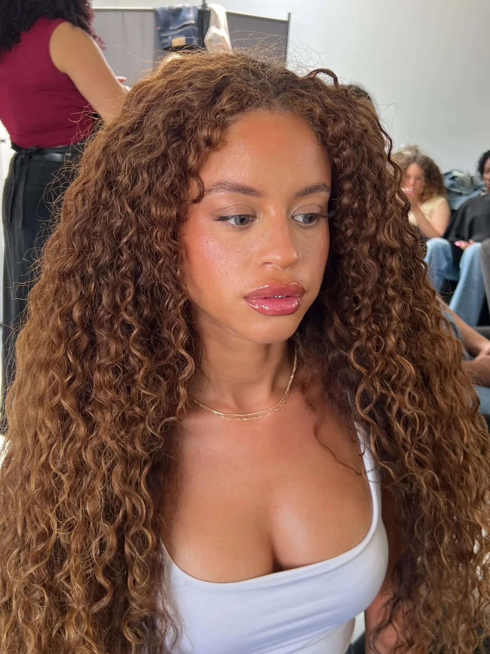 Jeune femme avec de longues boucles brunes clair assise dans une salle de maquillage en attente, entourée d'autres personnes.