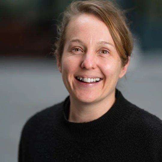 A woman with light brown hair smiling, wearing a black sweater, outdoors with a blurred background.
