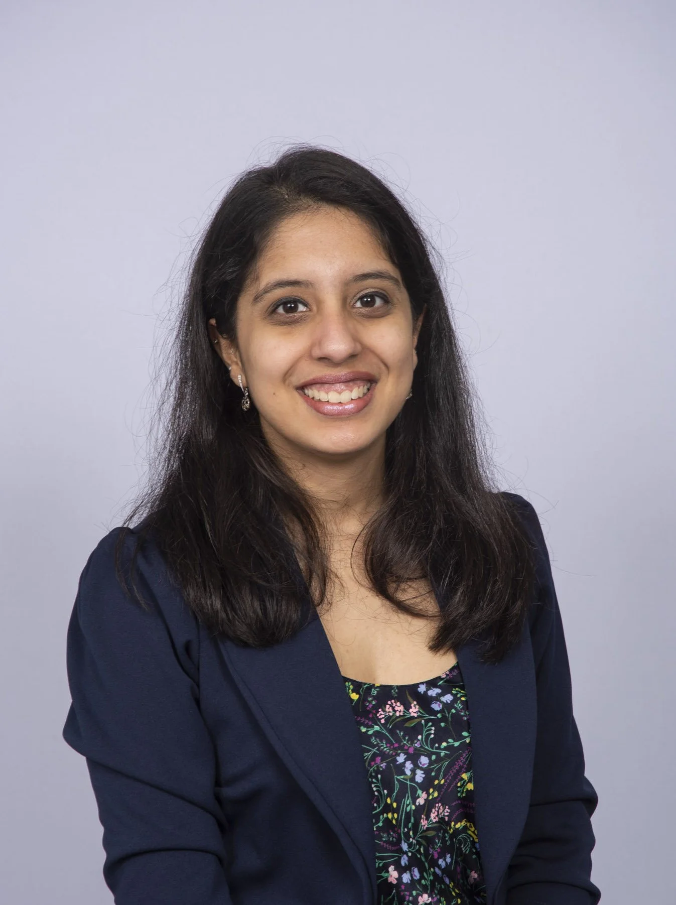 Portrait of a woman with shoulder-length black hair, wearing a dark blazer over a floral top, smiling against a light gray background.