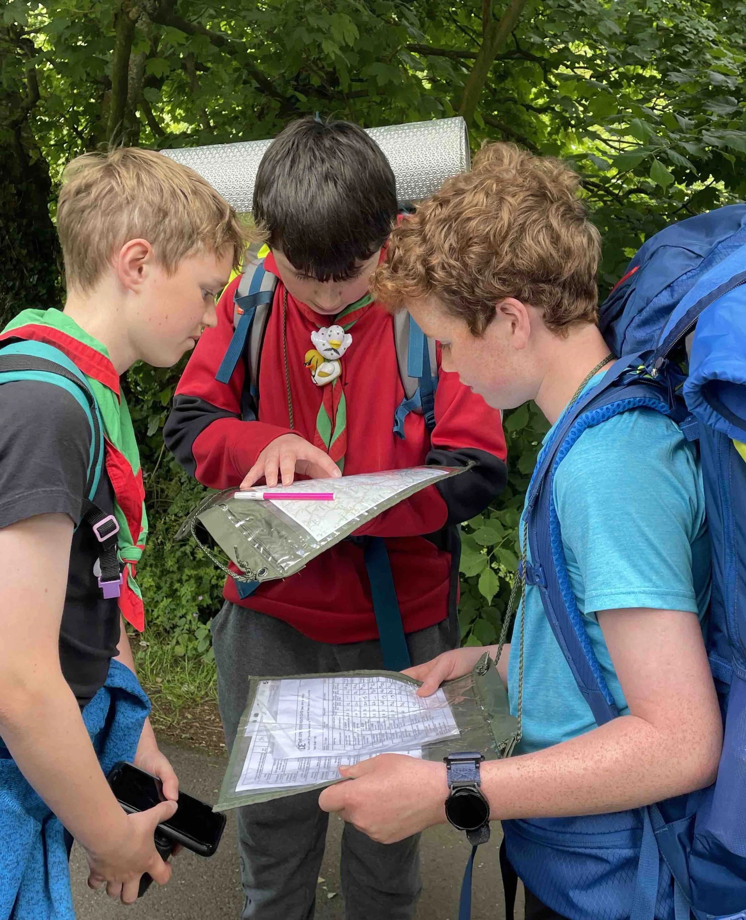 Three young boys with backpacks looking at a map with guidance from an older boy holding a plastic folder with maps, in a green outdoor setting.