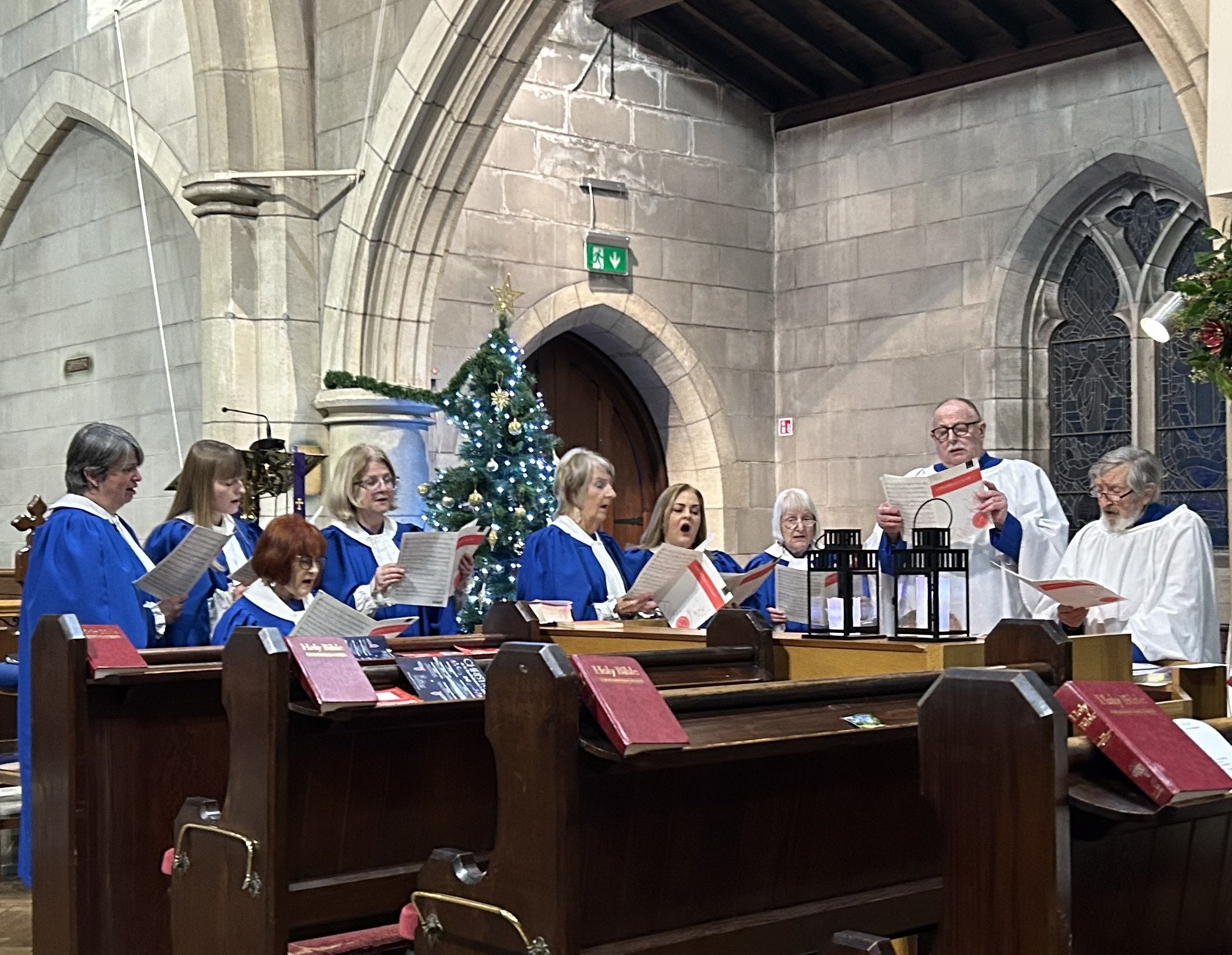 A church choir dressed in blue robes performing during a Christmas service. A decorated Christmas tree with lights and ornaments is visible behind them, and stained glass windows are on the wall. The choir members are holding songbooks and singing, with a conductor leading.
