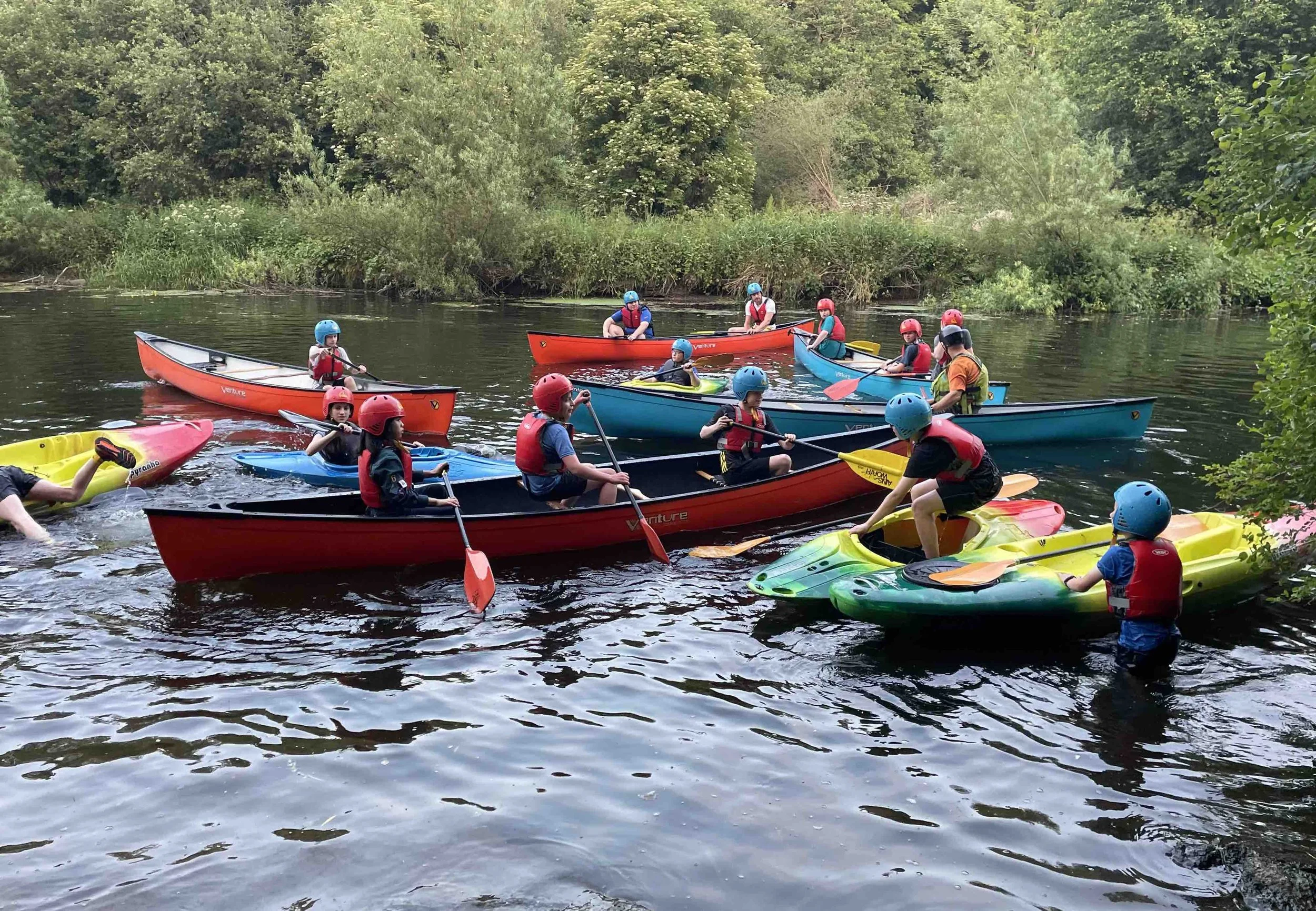 Group of people enjoying a kayaking outing on a river, with some in kayaks and others on paddleboards, all wearing helmets and life jackets amidst lush green trees.