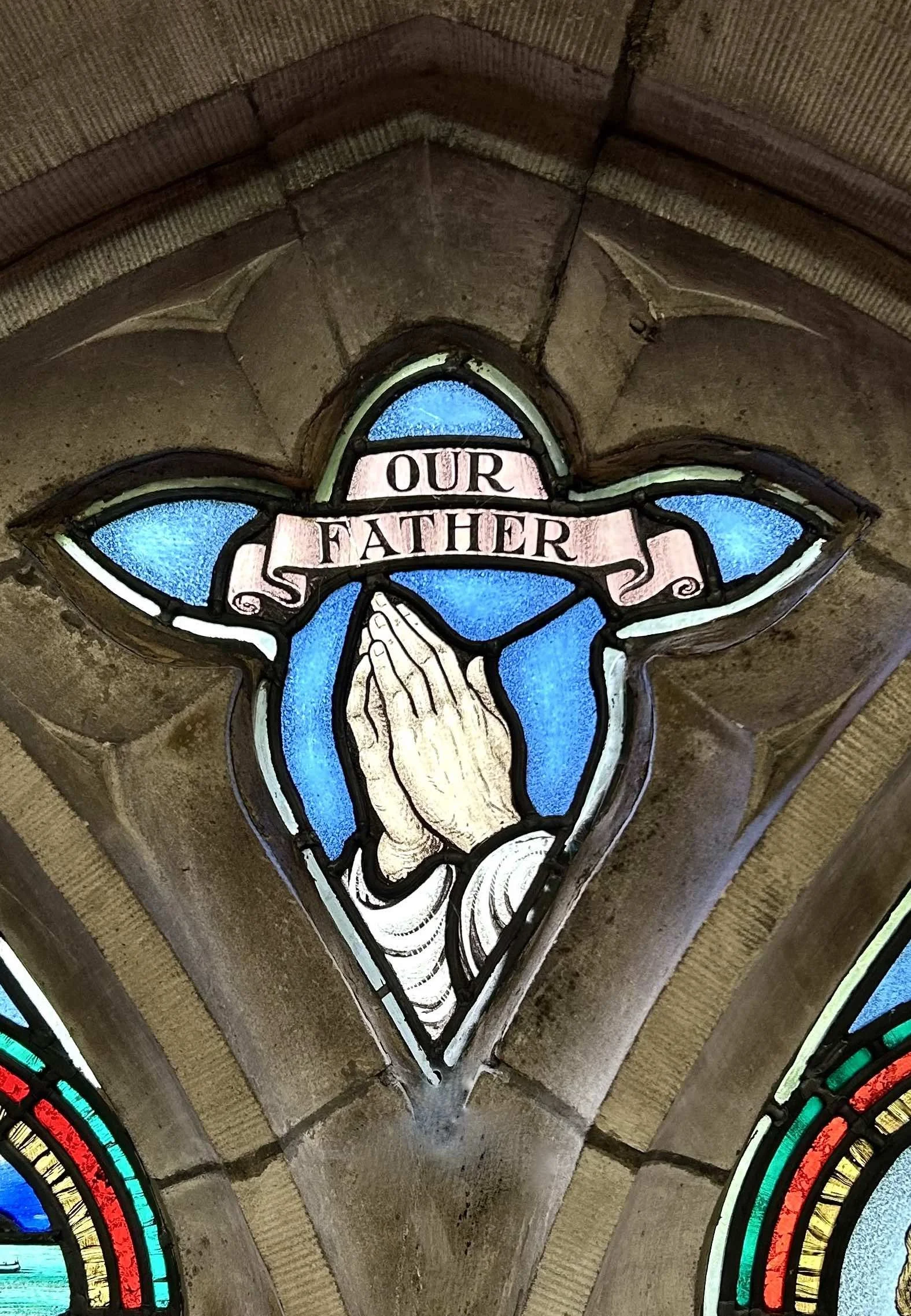 Stained glass window depicting praying hands with pink banner stating "Our Father" above, blue background, inside a stone church ceiling.
