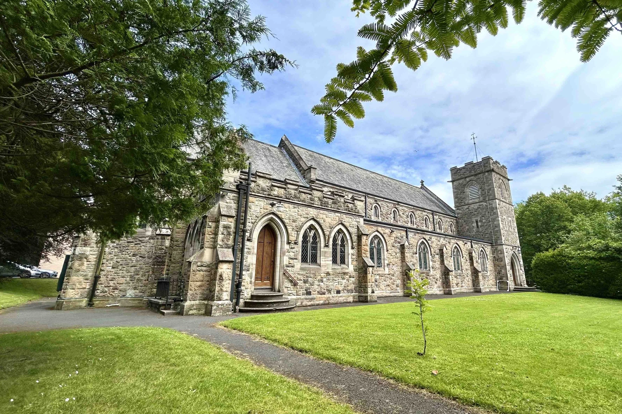 A stone church with arched windows and a tower, surrounded by green grass and trees, under a partly cloudy sky.