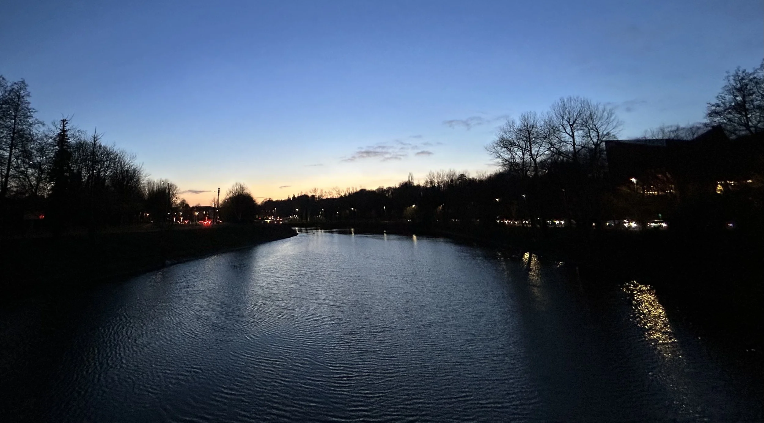 A river at dusk with a clear sky fading from sunset hues to dark blue, flanked by silhouetted trees and urban lights reflecting on the water.