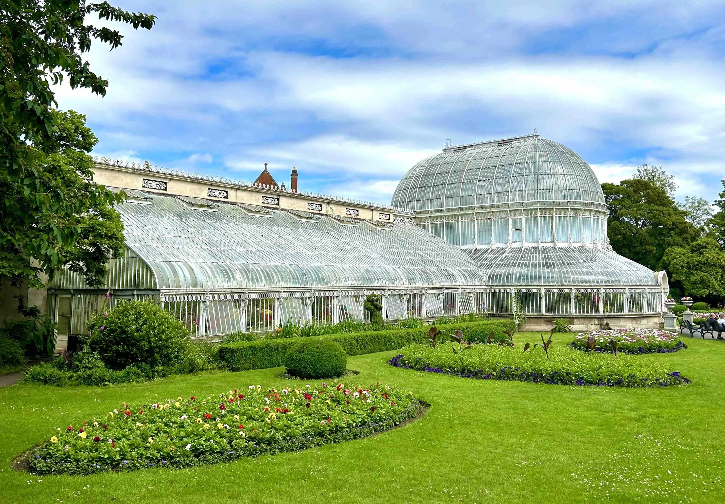 Glass greenhouse with a curved roof and domed top in a well-maintained garden with colorful flower beds and lush green grass, under a partly cloudy sky.
