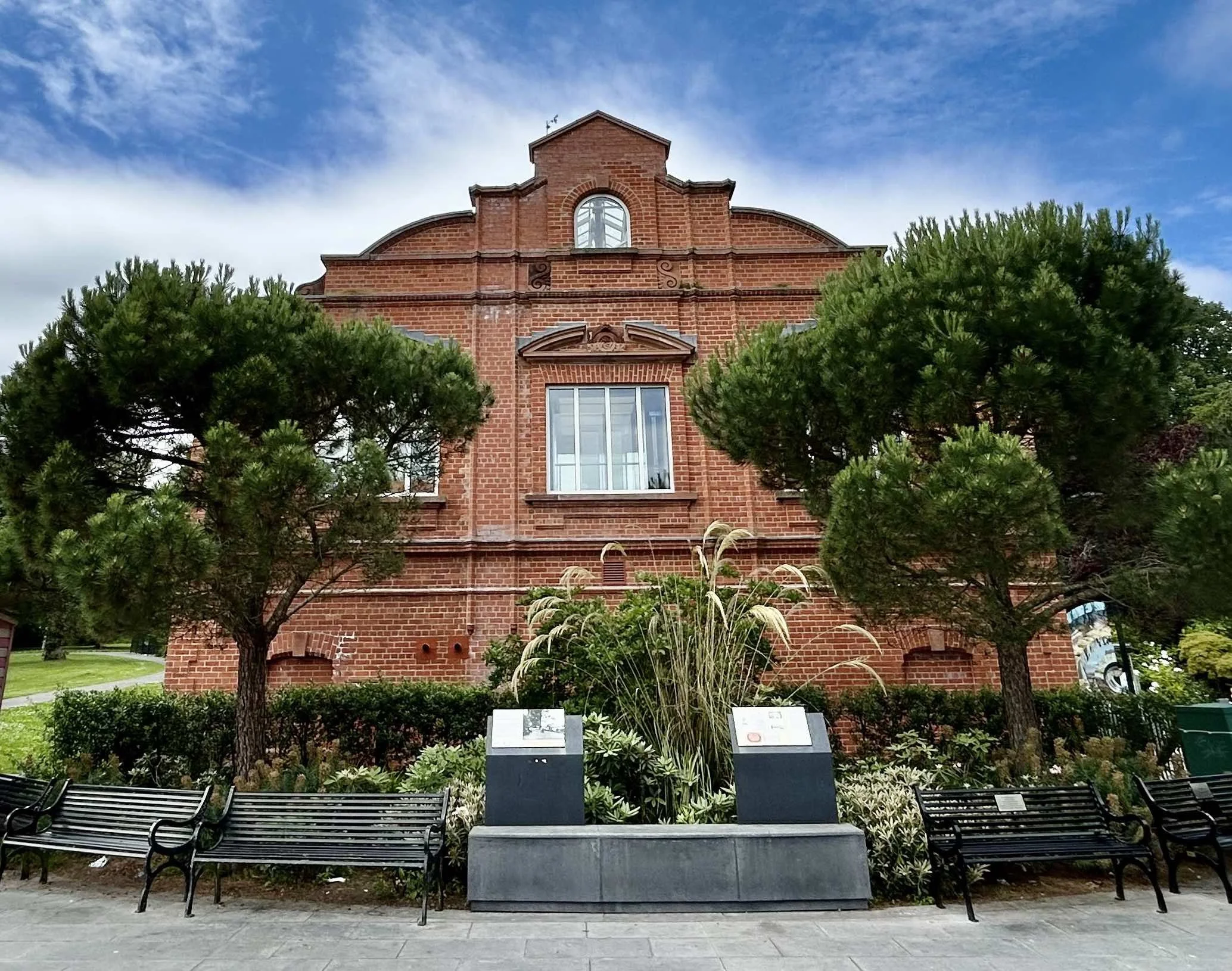Red brick building with two large trees in front, surrounded by benches and benches with informational plaques, under a partly cloudy sky.