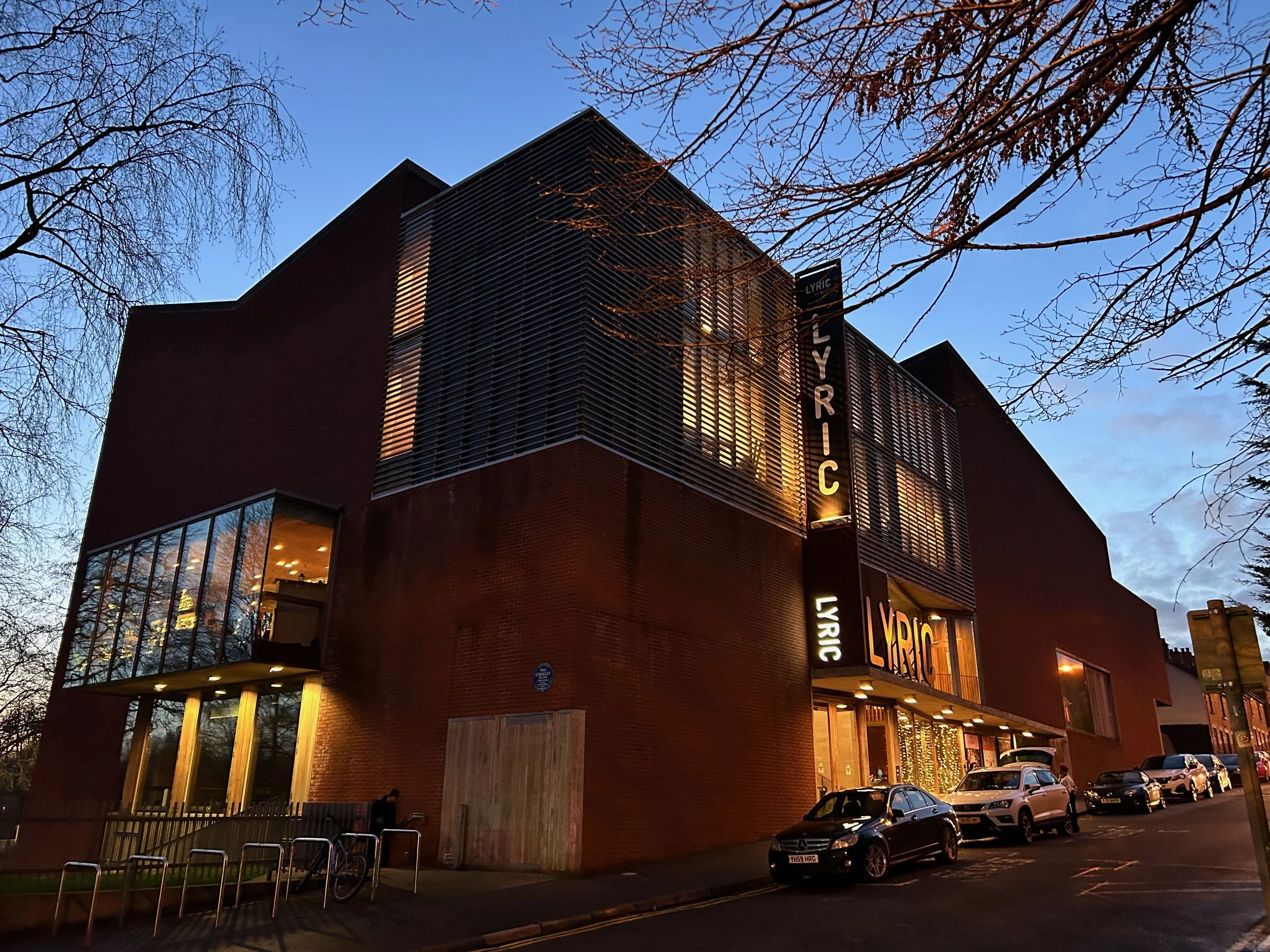 The Lyric building is a modern brick structure with large windows and illuminated vertical signage reading "Lyric" at dusk, with several parked cars outside and leafless trees framing the scene.