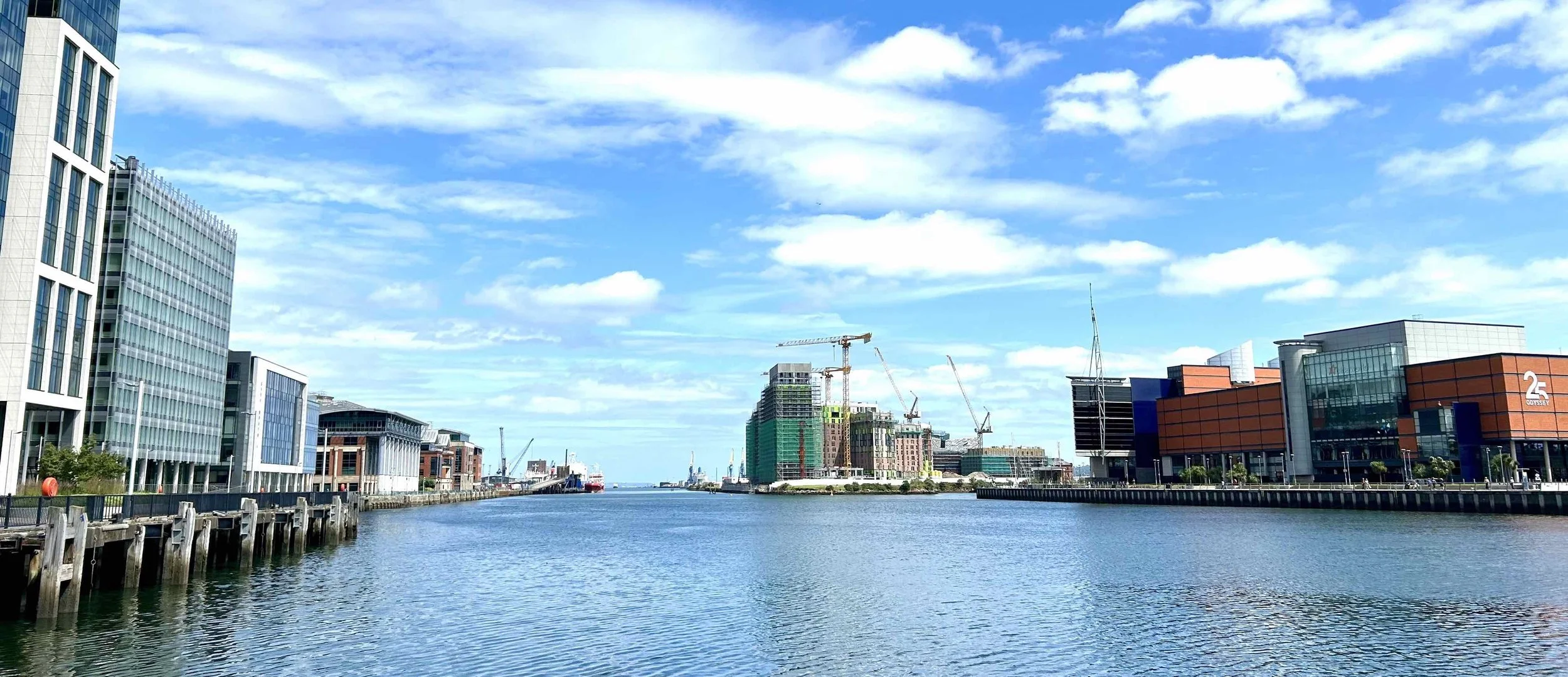 City waterfront with modern buildings under a partly cloudy sky, including construction cranes and water in the foreground.