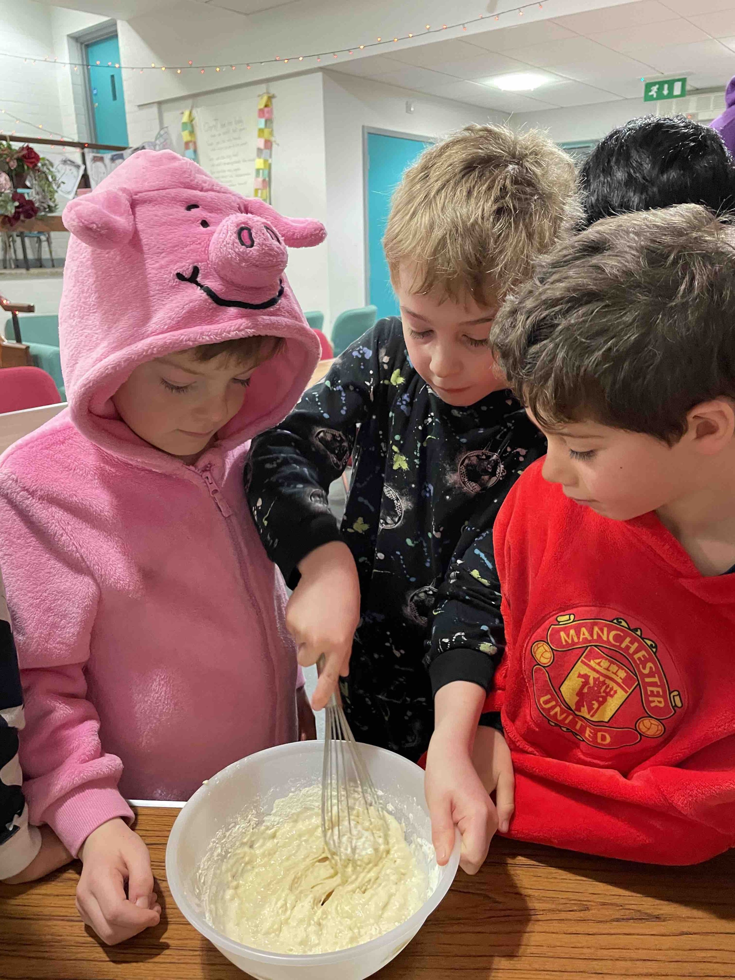 Three children, one in a pink Peppa Pig hoodie, are stirring a mixture in a white bowl at a wooden table. They are focused on the task.