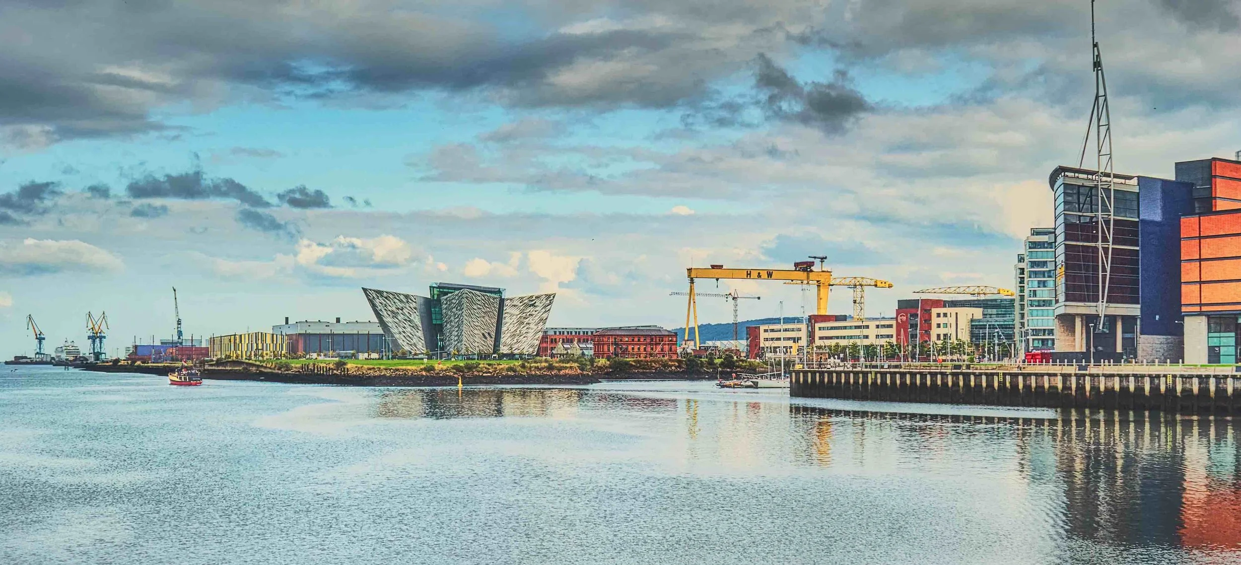 City skyline with modern buildings, cranes, and a body of water in the foreground, under a partly cloudy sky.