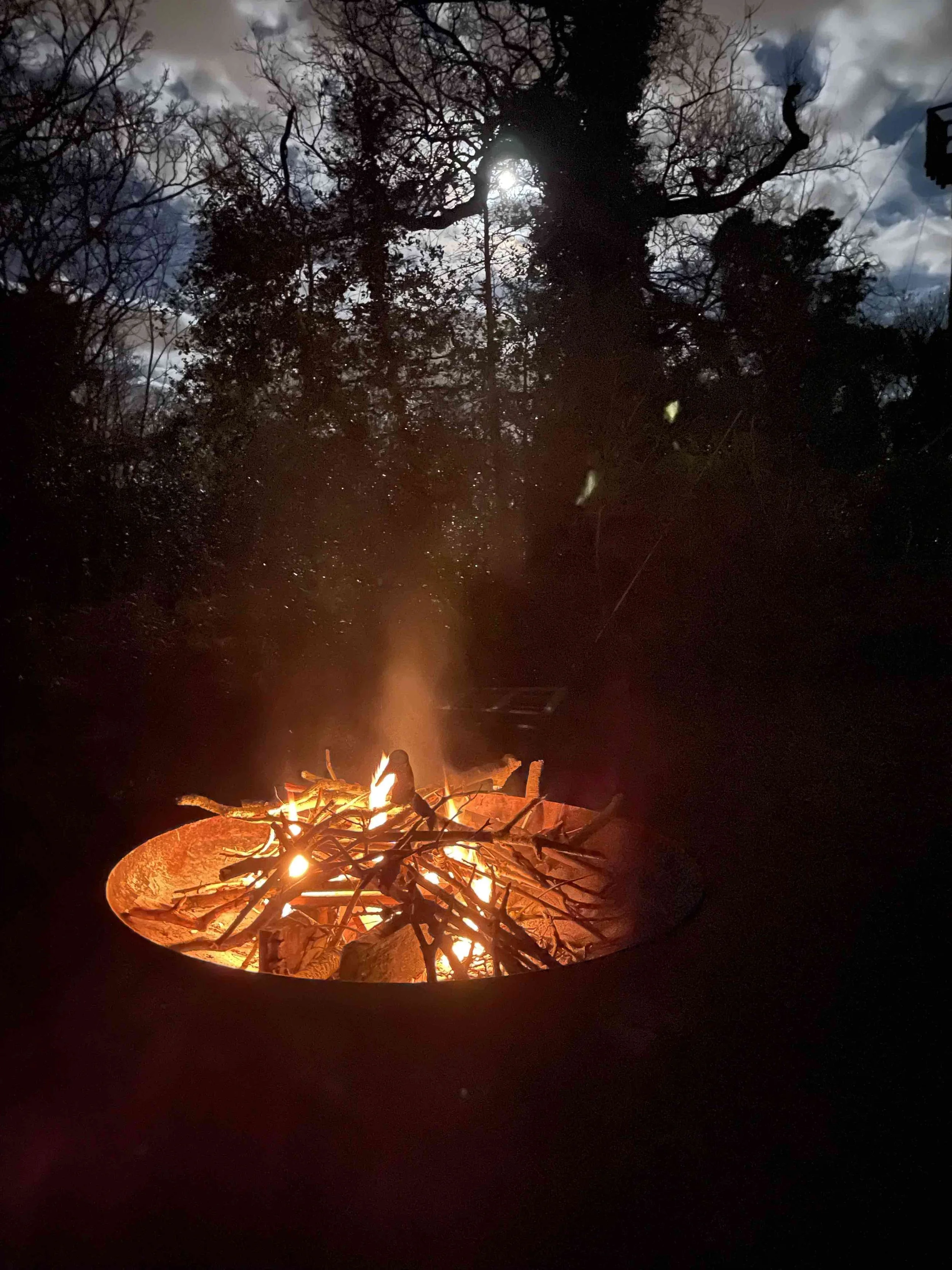 A campfire burning in a fire pit outdoors at night, with tree silhouettes and a cloudy sky with the moon overhead.