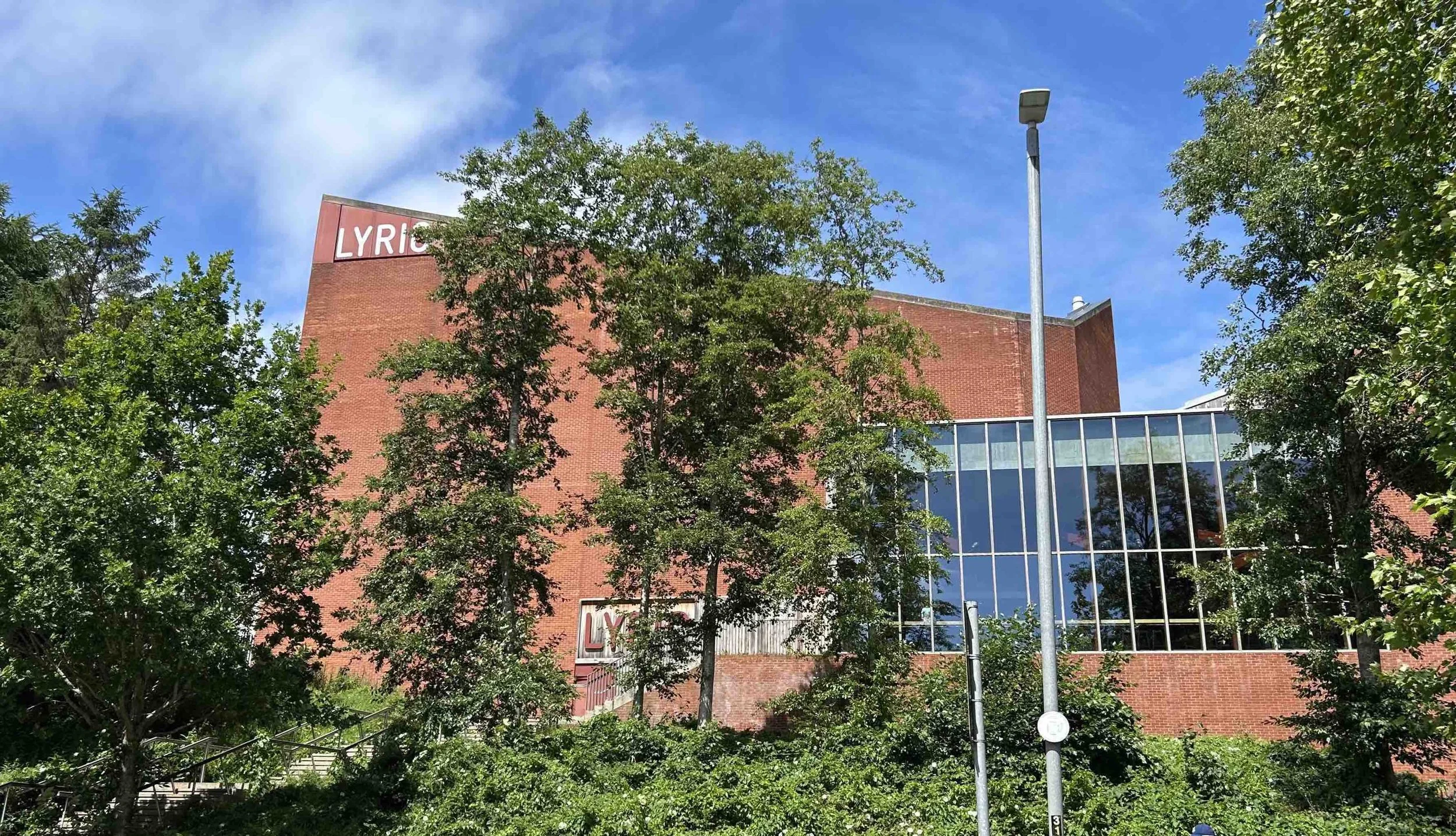 The Lyric Theatre Belfast. A red-brick building with the word "LYRIC" on it, partially obscured by trees, and blue sky with some clouds overhead.