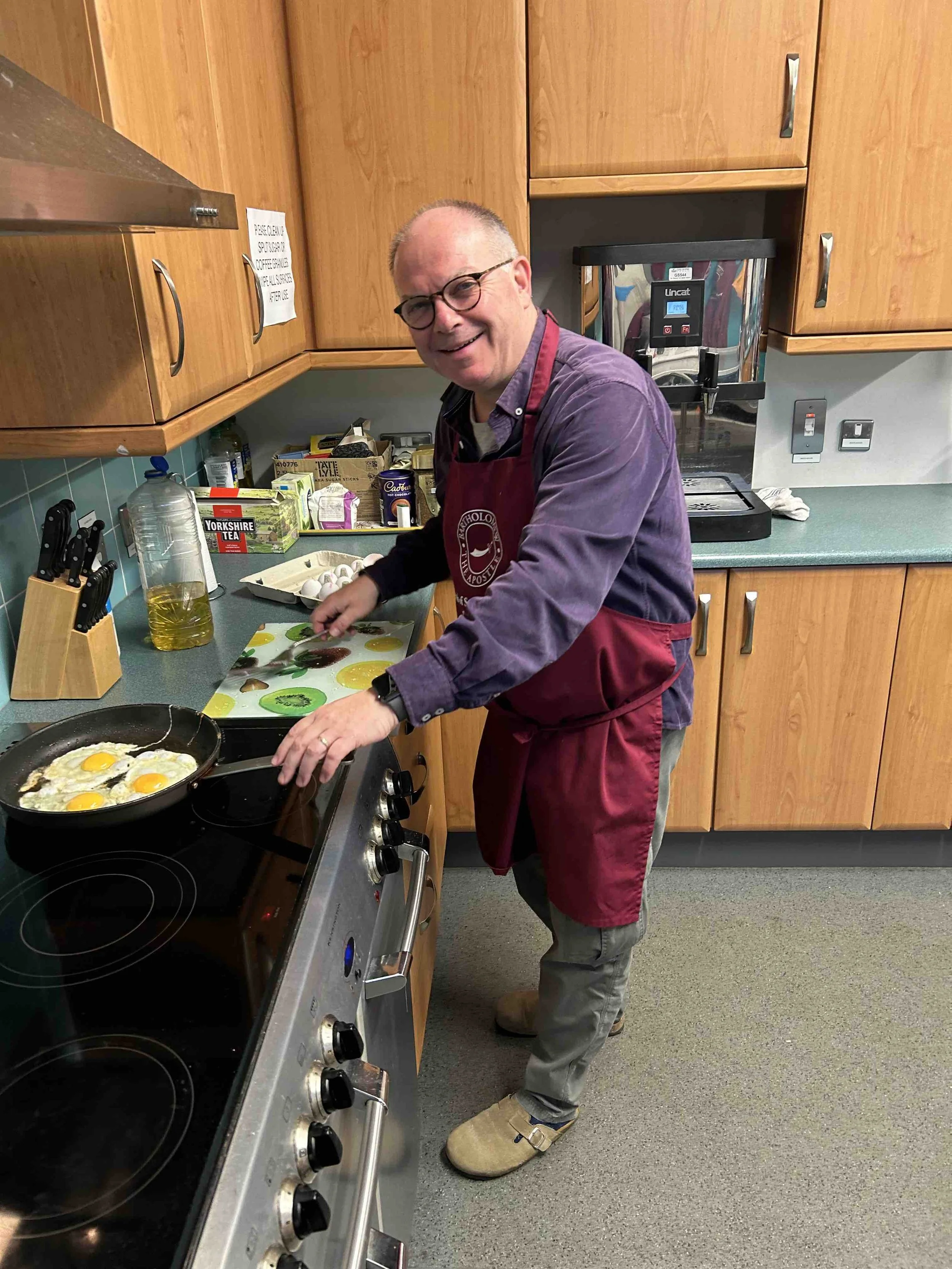 A man wearing glasses and a maroon apron cooking eggs in a frying pan in a kitchen.