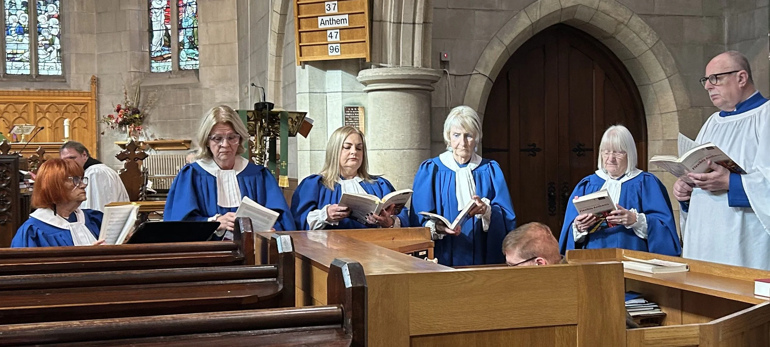 A choir of six women and one man standing and singing in a church, wearing blue and white choir robes. They are holding hymn books and appear to be engaged in a religious service. The church interior features stained glass windows, wooden pews, and stone walls.
