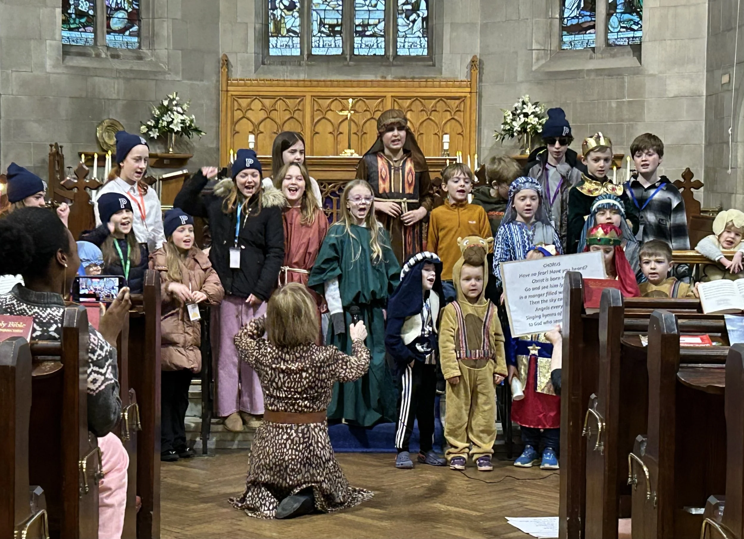 Children dressed in costumes performing a Christmas nativity scene in a church, with stained glass windows in the background.