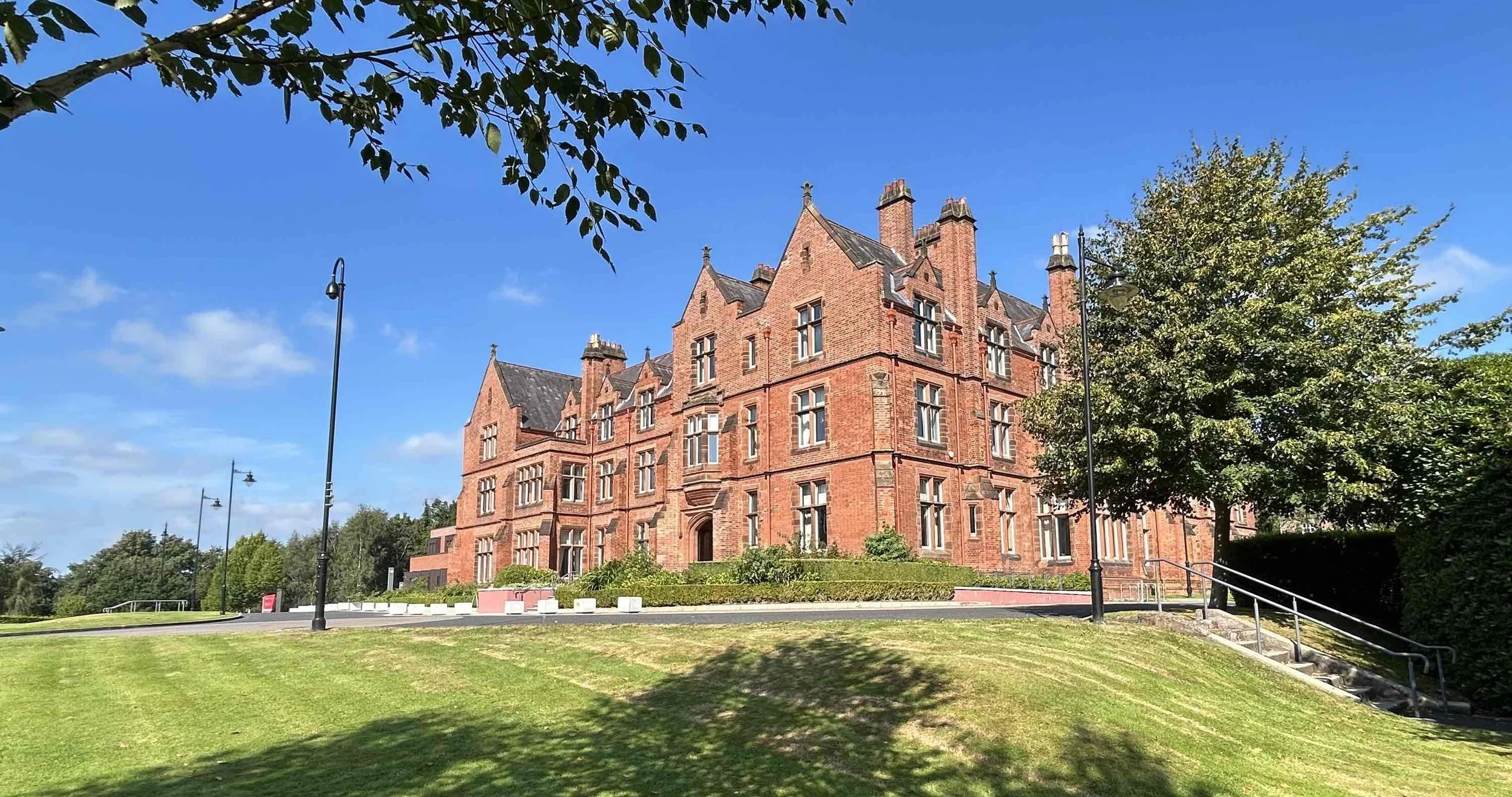 A large, historic red brick building with multiple gabled roofs and tall chimneys, surrounded by a well-maintained lawn and trees under a bright blue sky.
