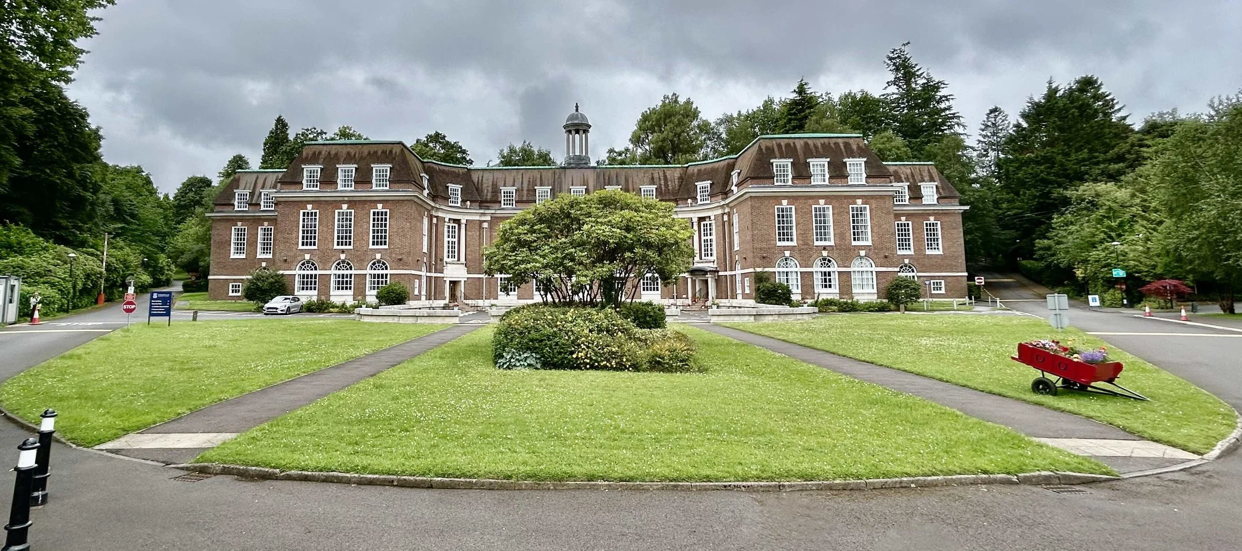 A large red brick building with numerous white-windowed floors, surrounded by a green lawn and trees, under a cloudy sky.
