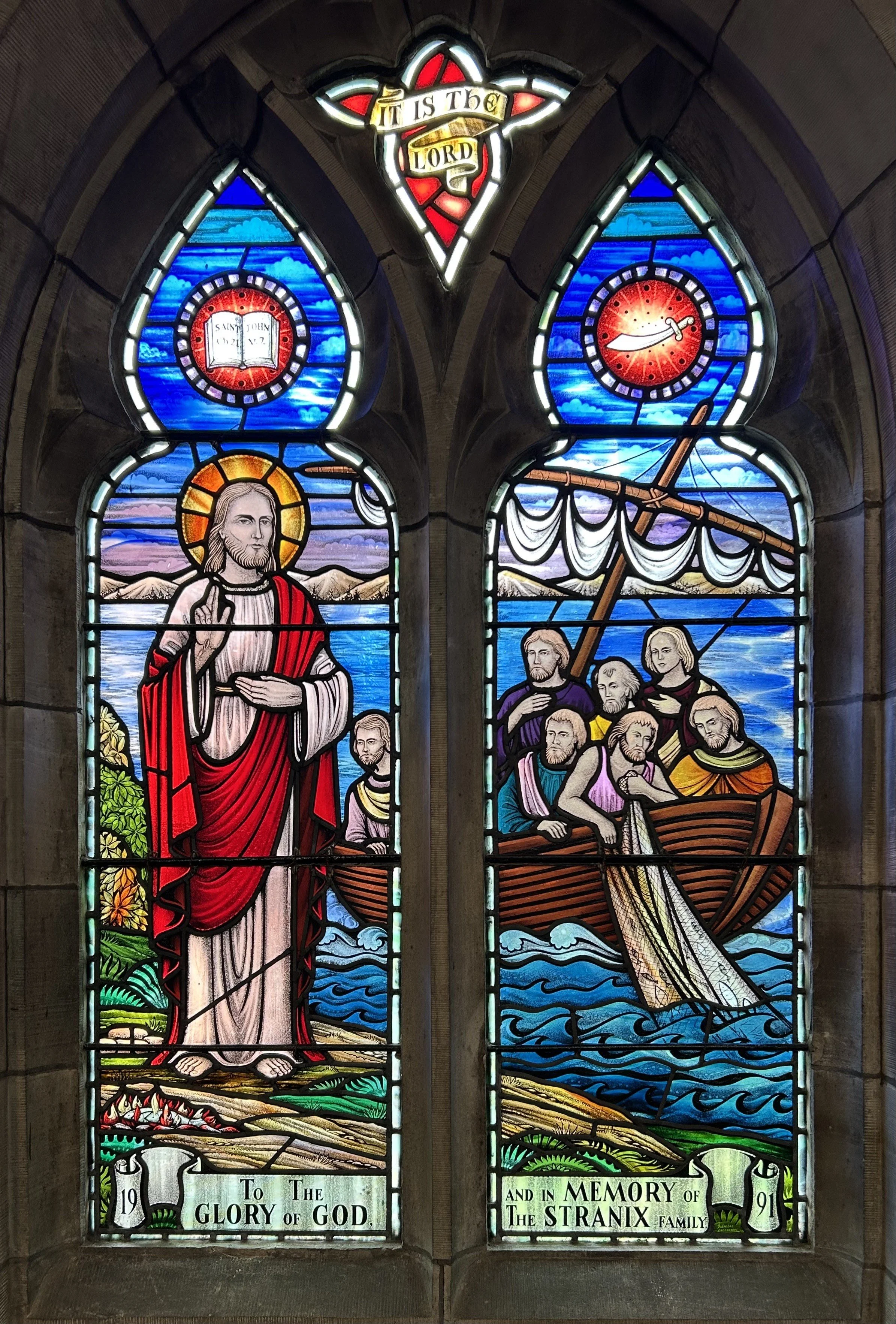 Colorful stained glass window depicting Jesus Christ standing near a lakeshore with mountains in the background, and seven men in a boat with a sailing ship's sail, under a sky with clouds and a clock. The top features a red heart with a sword and a banner reading 'It is the Lord'. The bottom inscriptions read 'To The Glory of God' and 'And In Memory of The Stranix Family', dated 1991.