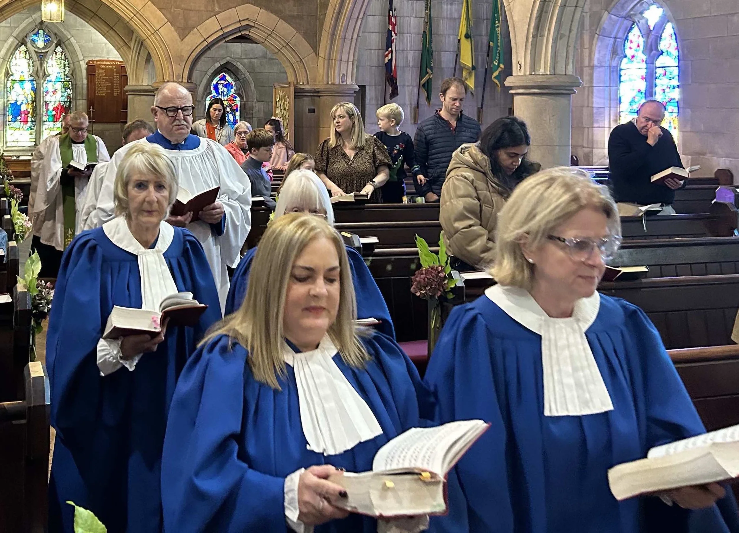 People participating in a church service, with many dressed in choir robes, inside a church with stained glass windows and flags in the background.