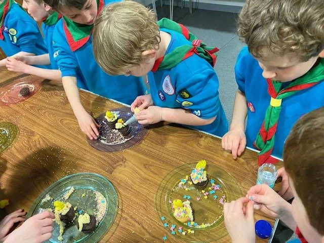 Group of children in blue uniforms and red scarves decorating cupcakes at a wooden table with colorful sprinkles and toppings.