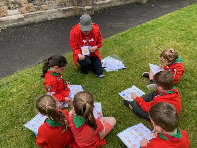 A woman and six children wearing red Scout uniforms with green neckerchiefs sitting on the grass outside, engaged in looking at picture books or pamphlets.