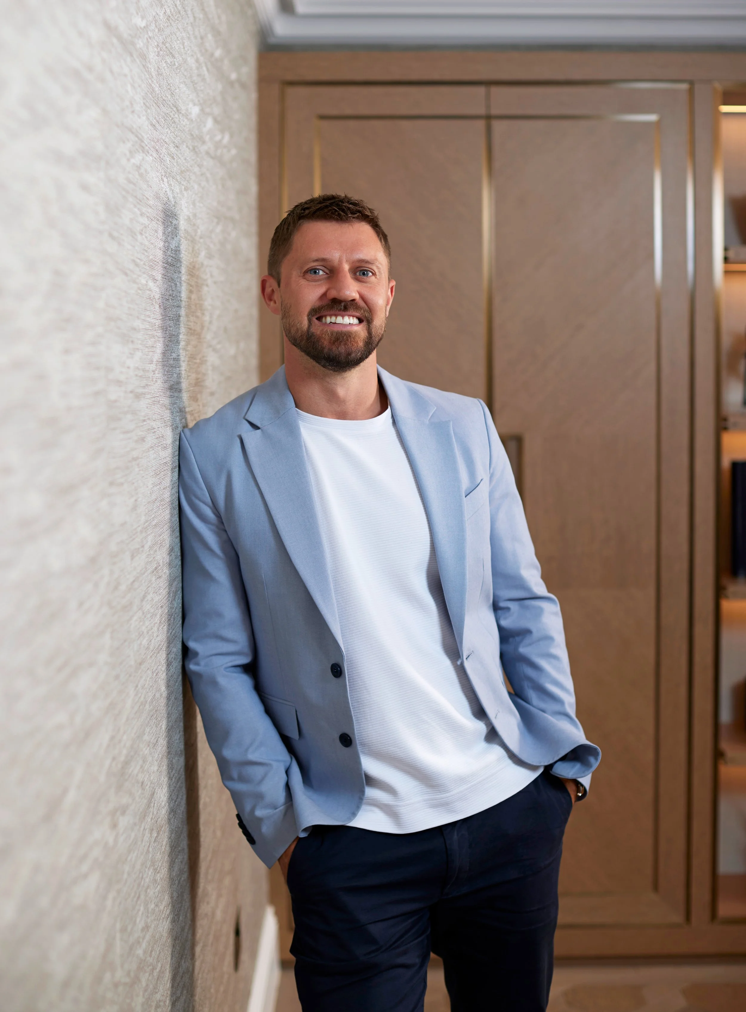A smiling man with a beard and short hair, dressed in a light blue blazer and white shirt, leaning against a textured wall indoors, with wooden shelving in the background.