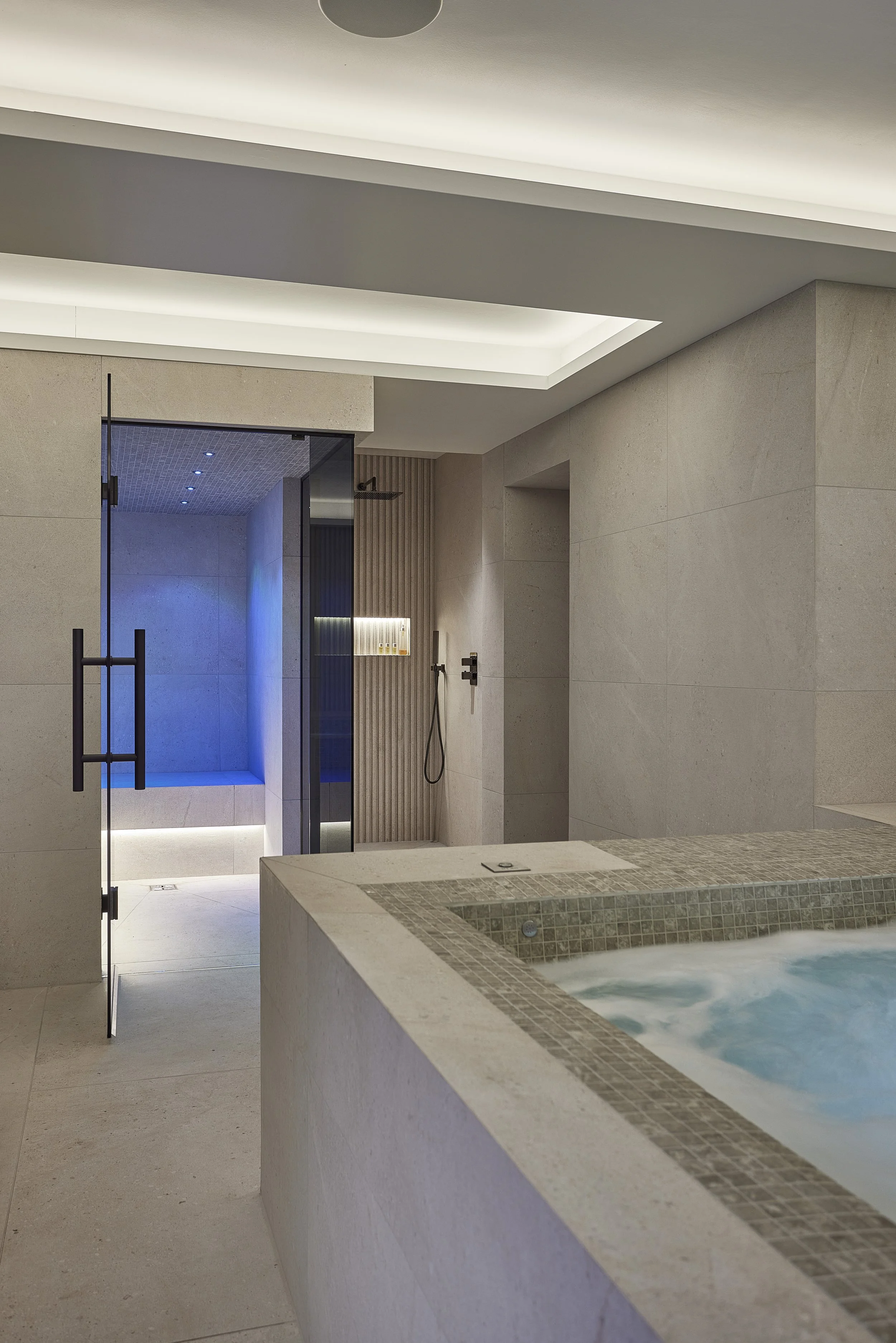 Modern indoor spa with a hot tub in the foreground, and a walk-in shower with a glass door in the background, featuring beige tiles, a ceiling-lit ceiling, and minimalist decor.