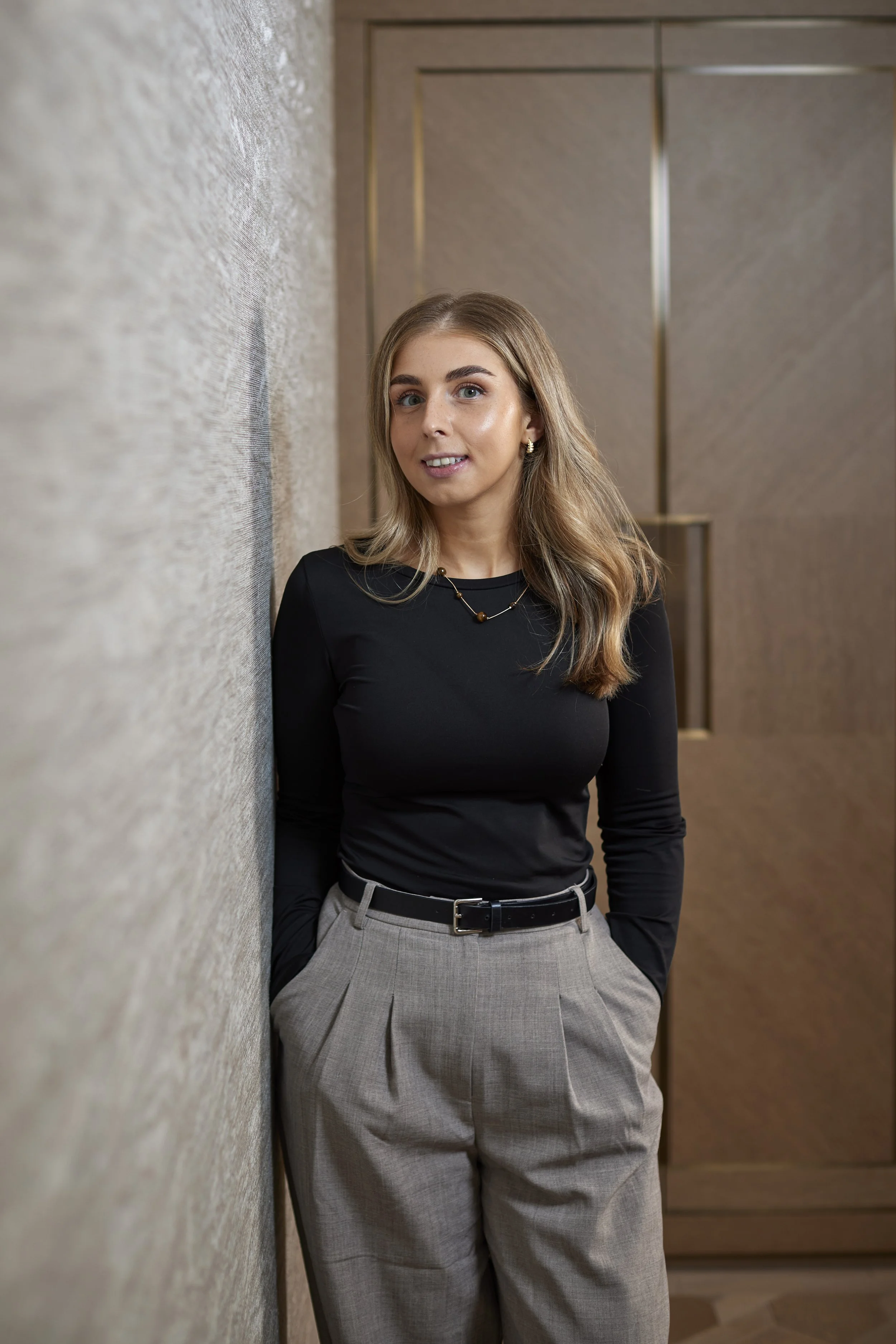 A young woman with blonde hair wearing a black long-sleeve top and beige high-waisted trousers stands next to a textured wall in an indoor setting with wooden paneling in the background.